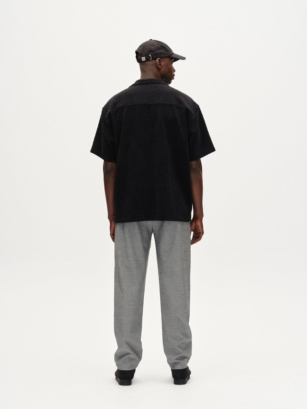 A man stands with his back to the camera, wearing the Gabba Jens Over Diam SS shirt in black, gray trousers, black shoes, and a black cap against a plain white background.