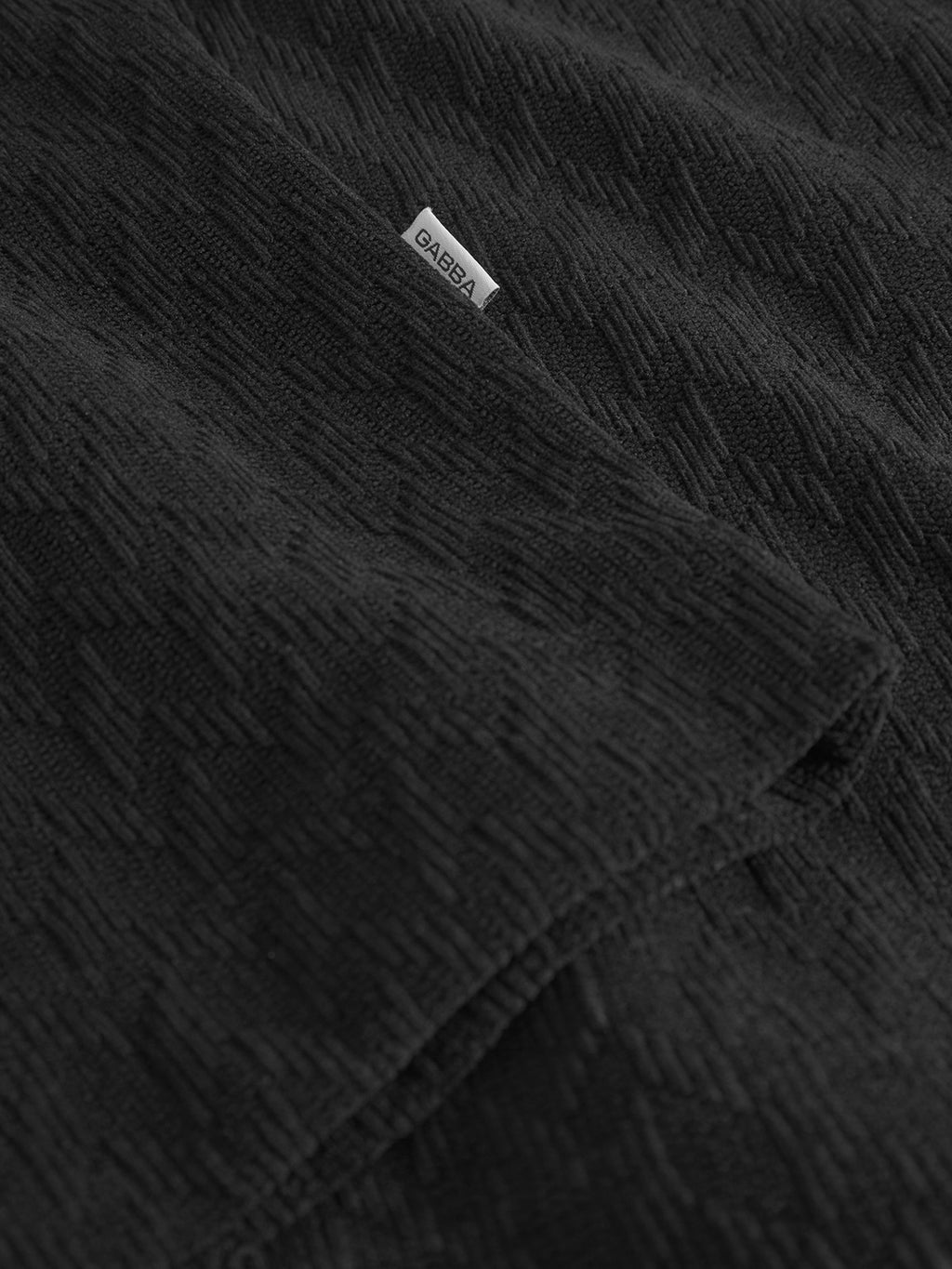 Close-up of the textured black Jens Over Diam SS shirt by Gabba, featuring diamond jacquard velvet and a subtle geometric pattern. A small white DABBA label is stitched on, perfect for an oversized resort look.