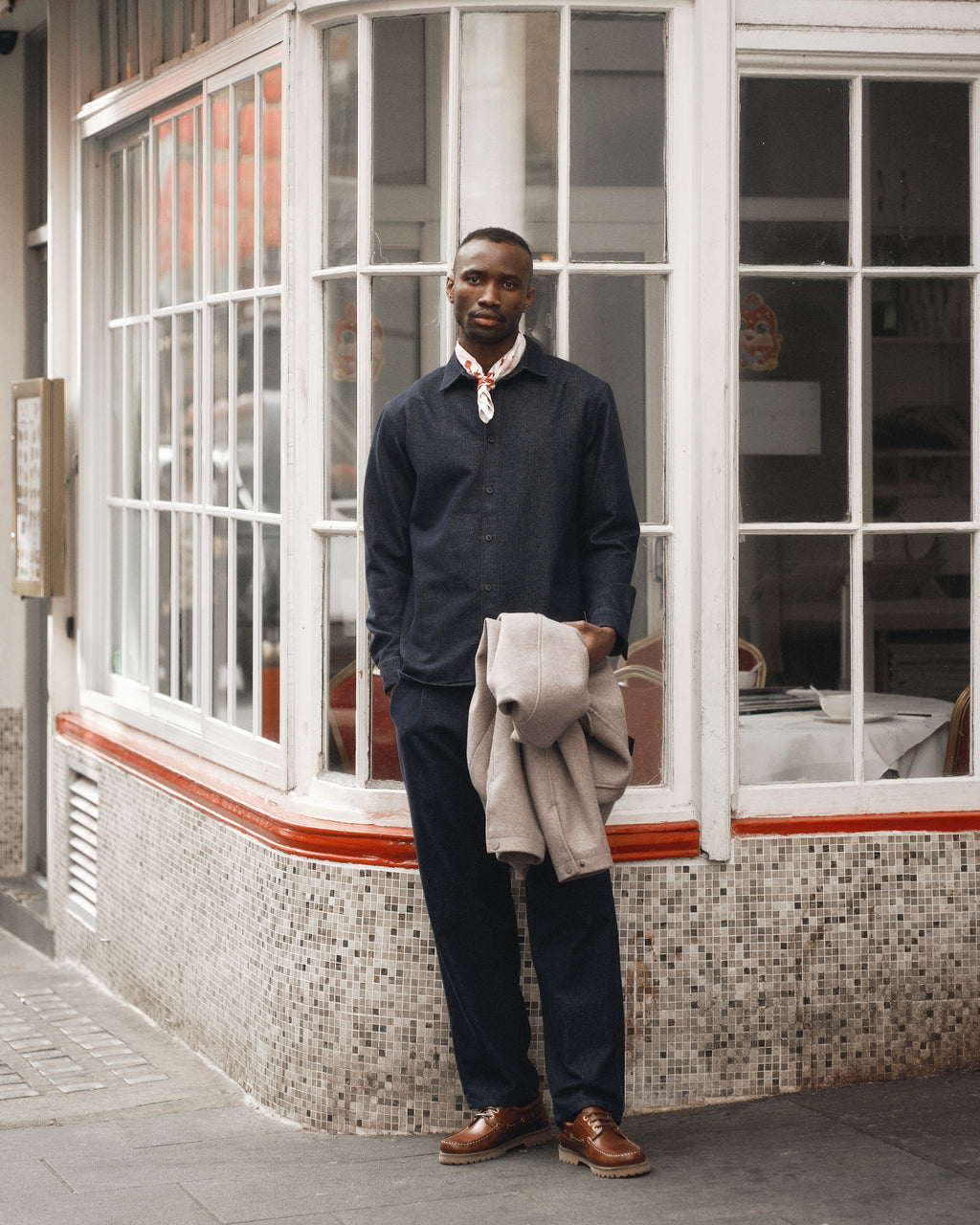 A man stands on a city sidewalk in front of large windows, wearing Gabbas David Chev - Dark Navy shirt with loose-tapered chinos, brown shoes, and a patterned neck scarf. He holds a light herringbone jacket and looks directly at the camera.