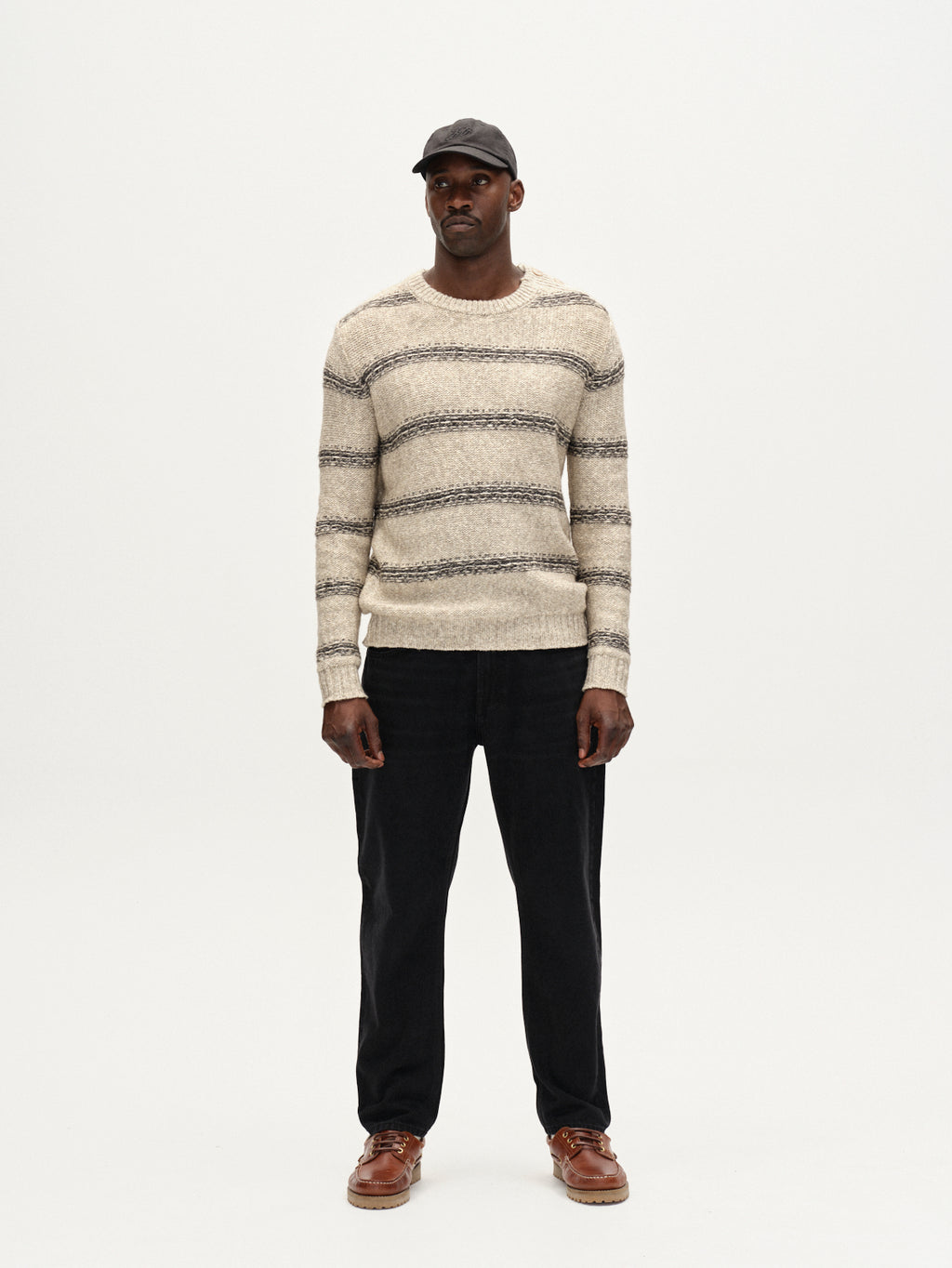 A man stands facing forward against a plain white background, wearing a dark cap, cream and gray striped sweater, brown shoes, and Gabbas Carl Dlak - Black Denim jeans.