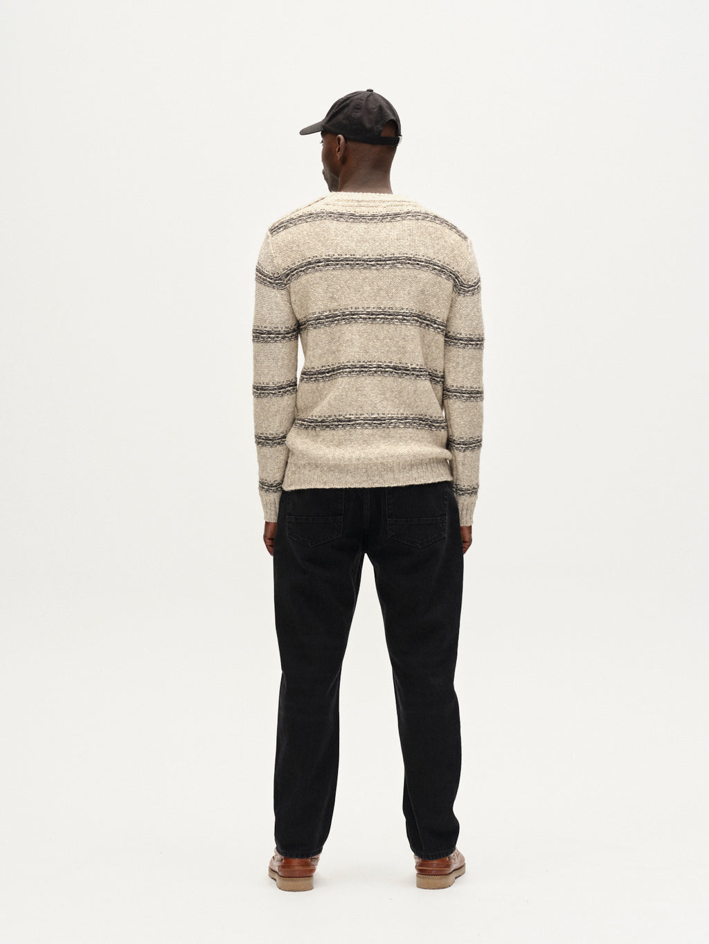A man faces away from the camera, wearing a black cap, beige and gray striped sweater, brown shoes, and Gabba Carl Dlak - Black Denim jeans against a plain white background.