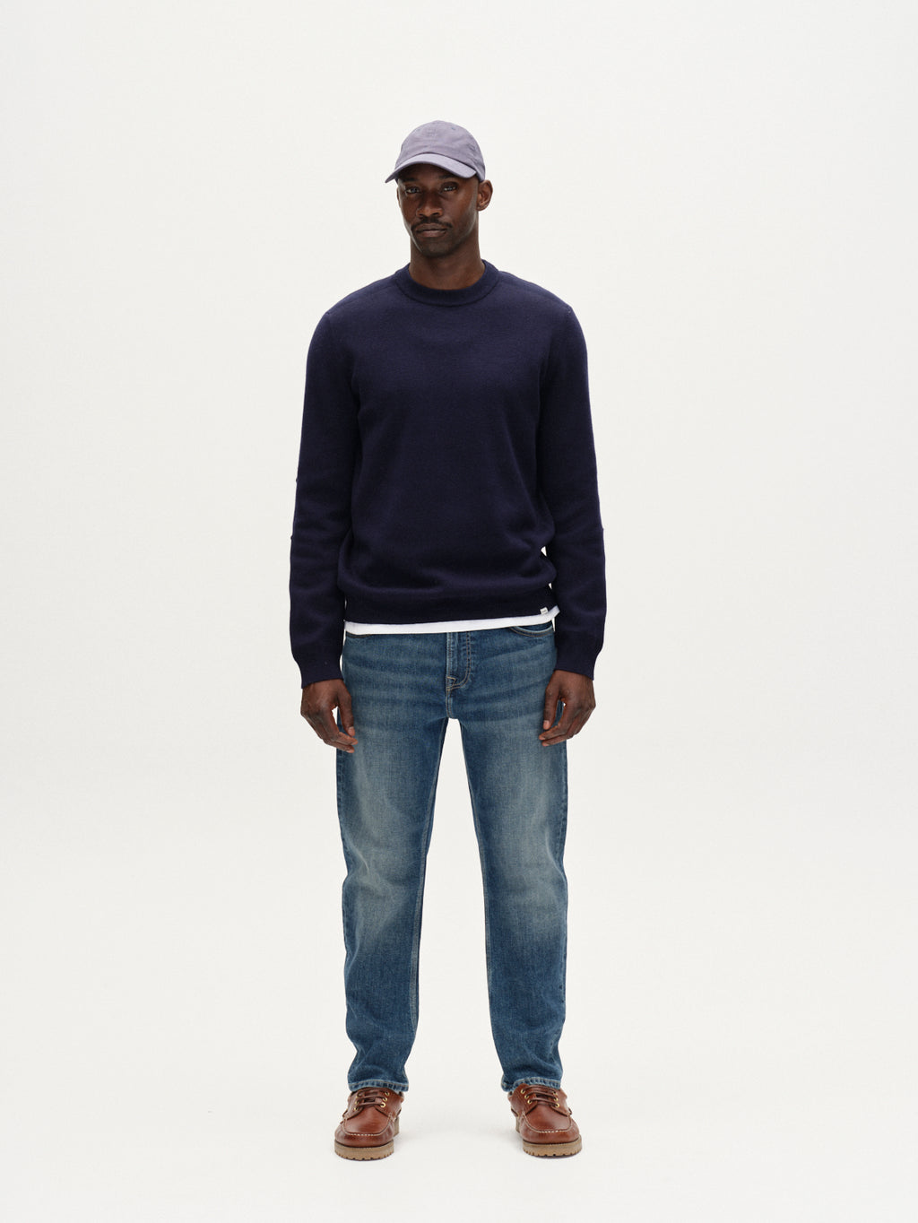 A man faces forward against a plain white background, wearing a light purple cap, navy sweater, Gabba Carl Jingo - Dark Blue Denim relaxed fit stretch jeans, and brown shoes.