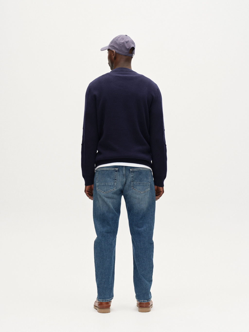 A man faces away from the camera, wearing Gabba Carl Jingo - Dark Blue Denim jeans, a navy sweater, brown shoes, and a light purple baseball cap against a plain white background.