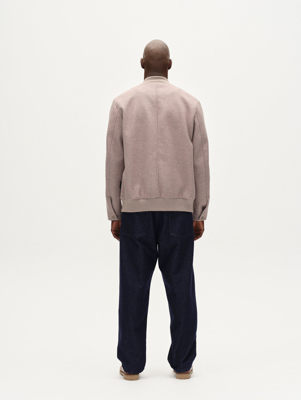 A person stands with their back to the camera, wearing the Gabba Orta Wox - Sand breathable bomber jacket, loose dark blue pants, and beige shoes in front of a plain white background.