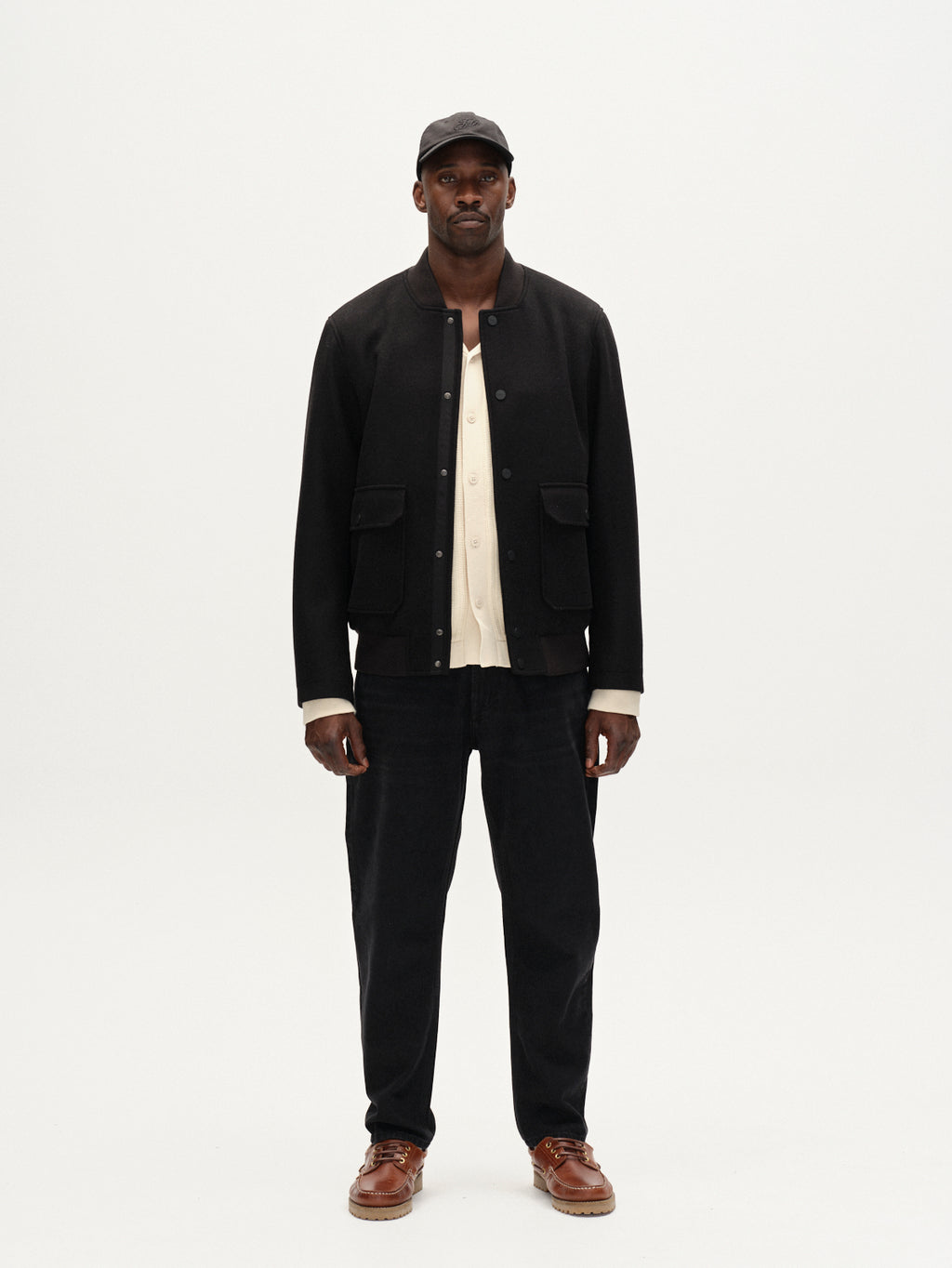 A man faces forward against a plain white background, wearing Gabba’s Orta Wox - Black windproof bomber jacket with front pockets, paired with a black cap, cream shirt, black pants, and brown shoes.