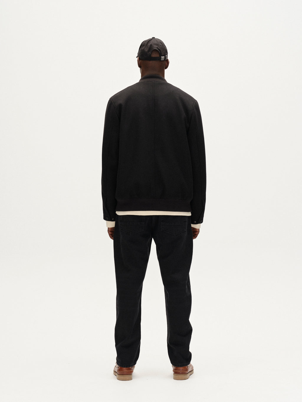 A man faces away from the camera, wearing the Gabba Orta Wox - Black windproof bomber jacket, black pants, a black cap, and brown shoes against a plain white background.
