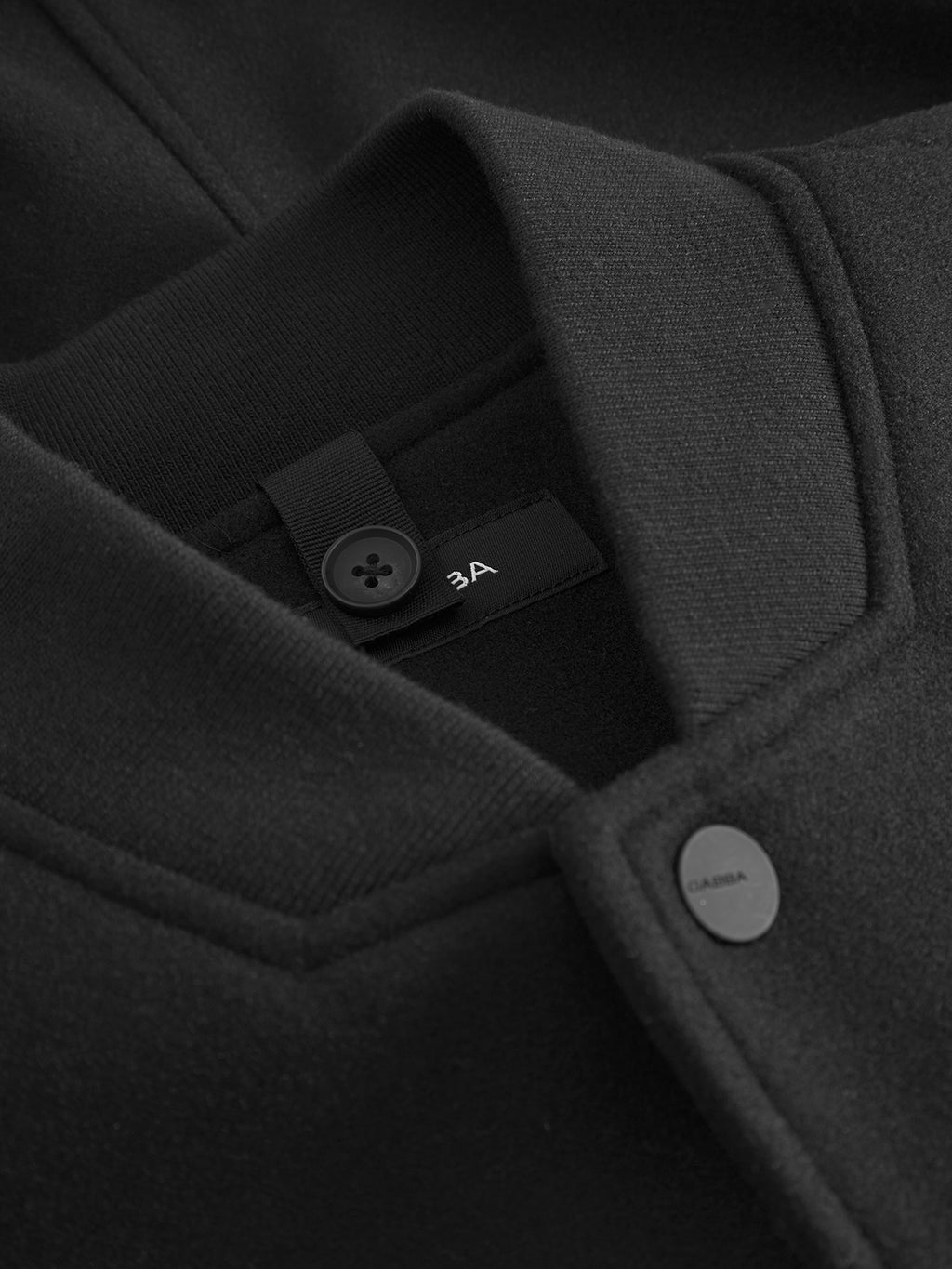 Close-up of Orta Wox - Black bomber jacket by Gabba shows its buttoned collar strap and a black label with 3A in white. The CARIMA-engraved snap highlights the windproof, performance fabric construction.