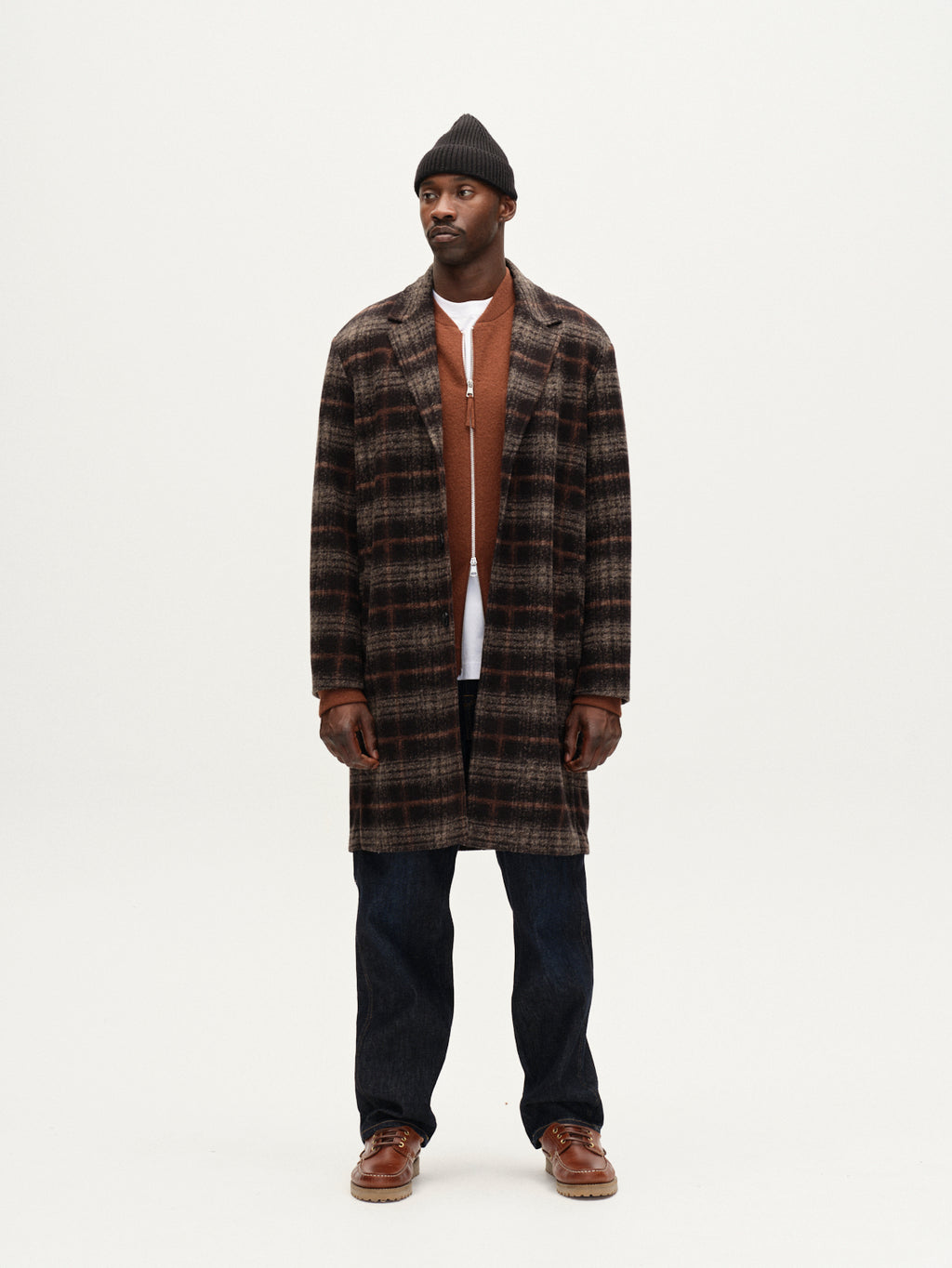 A man wearing the Gabba Leeds Cast - Multi Check brown plaid overcoat, a rust shirt over a white tee, black beanie, dark jeans, and brown shoes stands against a plain white background.