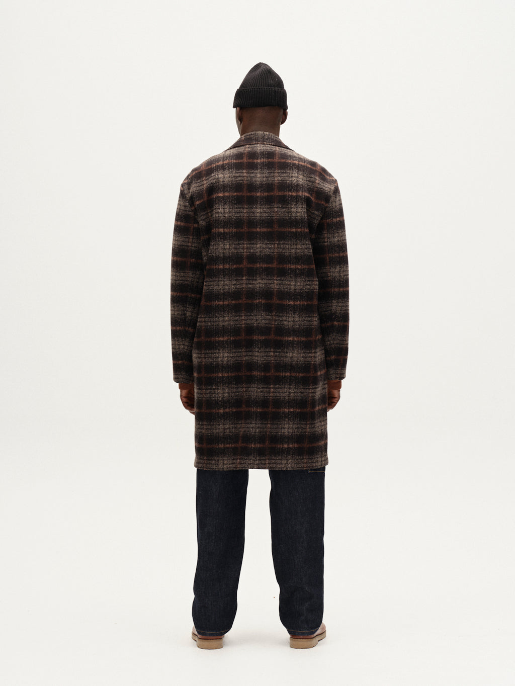 A person faces away, wearing Gabbas Leeds Cast - Multi Check coat—a long, oversized plaid in dark tones—paired with a black beanie, dark jeans, and light brown shoes against a plain white background.