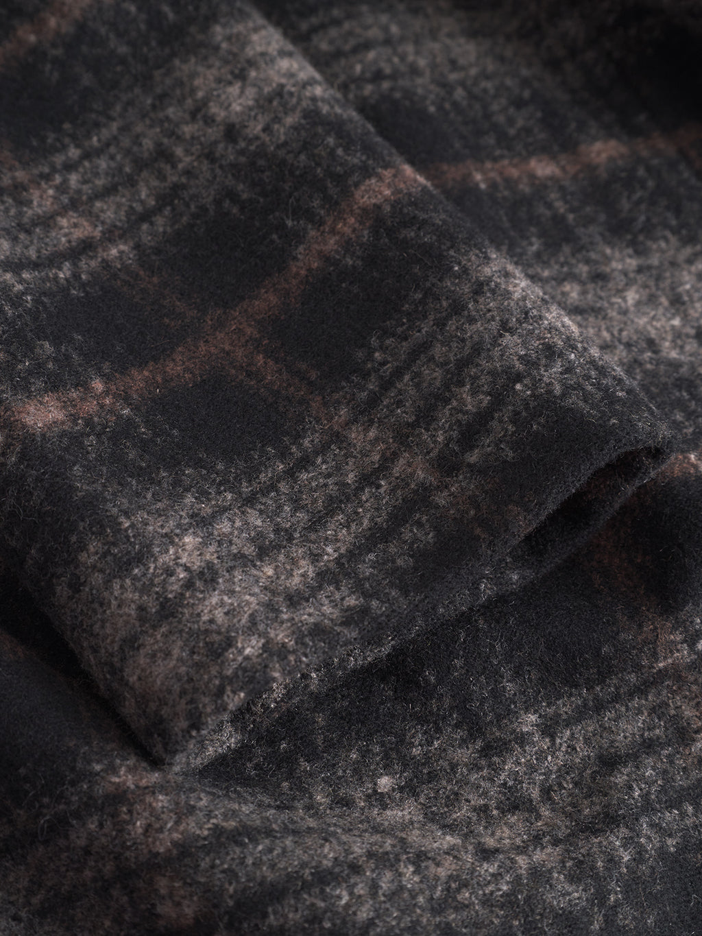 Close-up of Gabba’s Leeds Cast - Multi Check: a soft, brushed wool-blend fabric with a blurred winter check pattern in dark gray and muted brown tones. Its textured, slightly fuzzy surface shows gentle folds and subtle shadows.