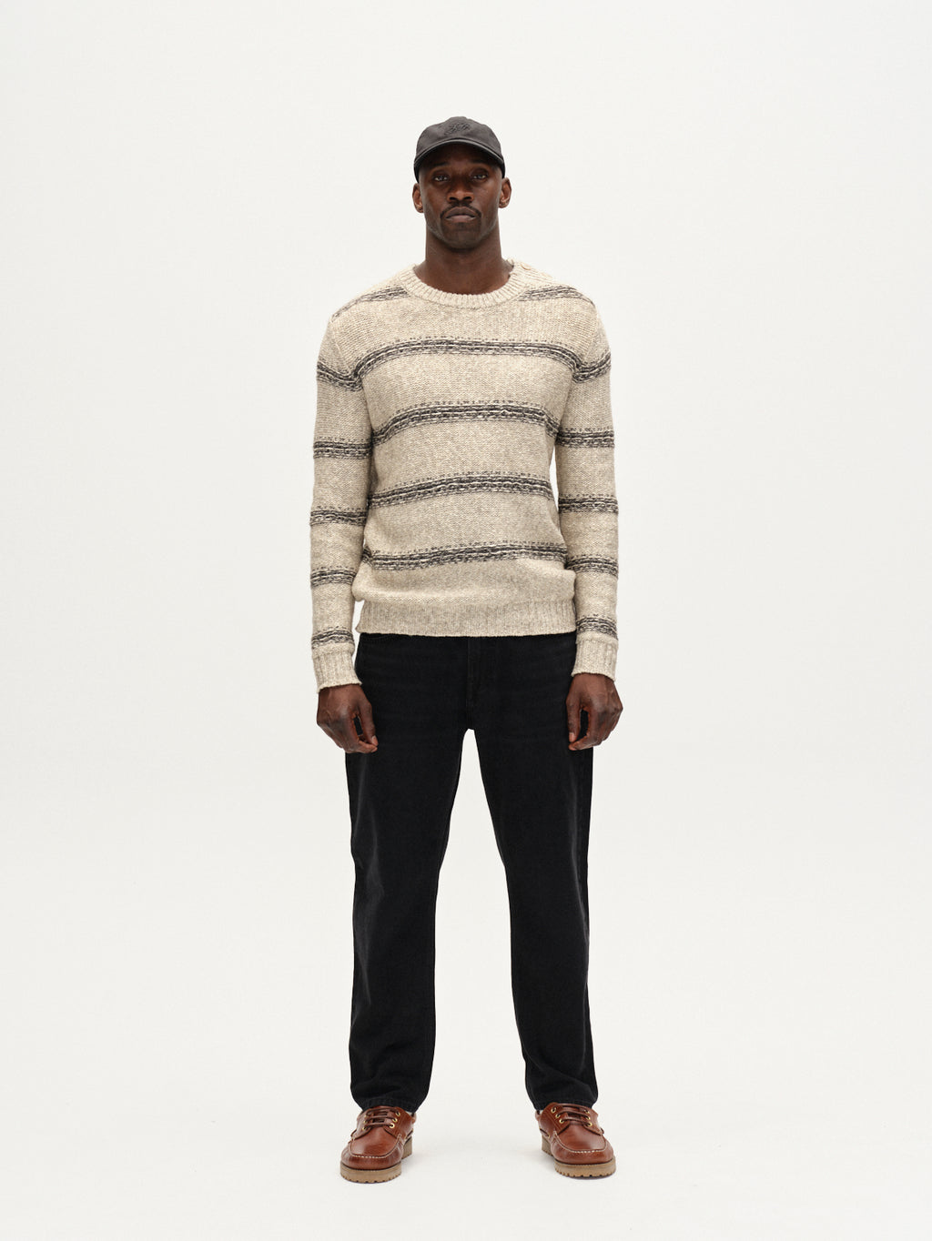 A man faces forward against a white background, wearing the Gabba Osaka Fay LS - Ecru striped long-sleeve top, black pants, brown shoes, and a black baseball cap.