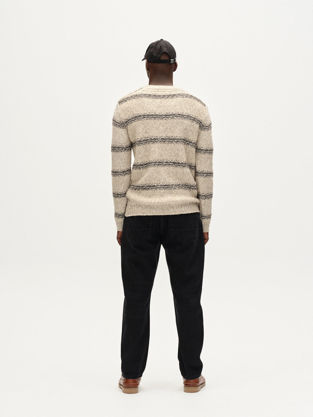 A person in the Gabba Osaka Fay LS - Ecru top, styled with black pants, brown shoes, and a black cap, stands facing away against a plain white background.