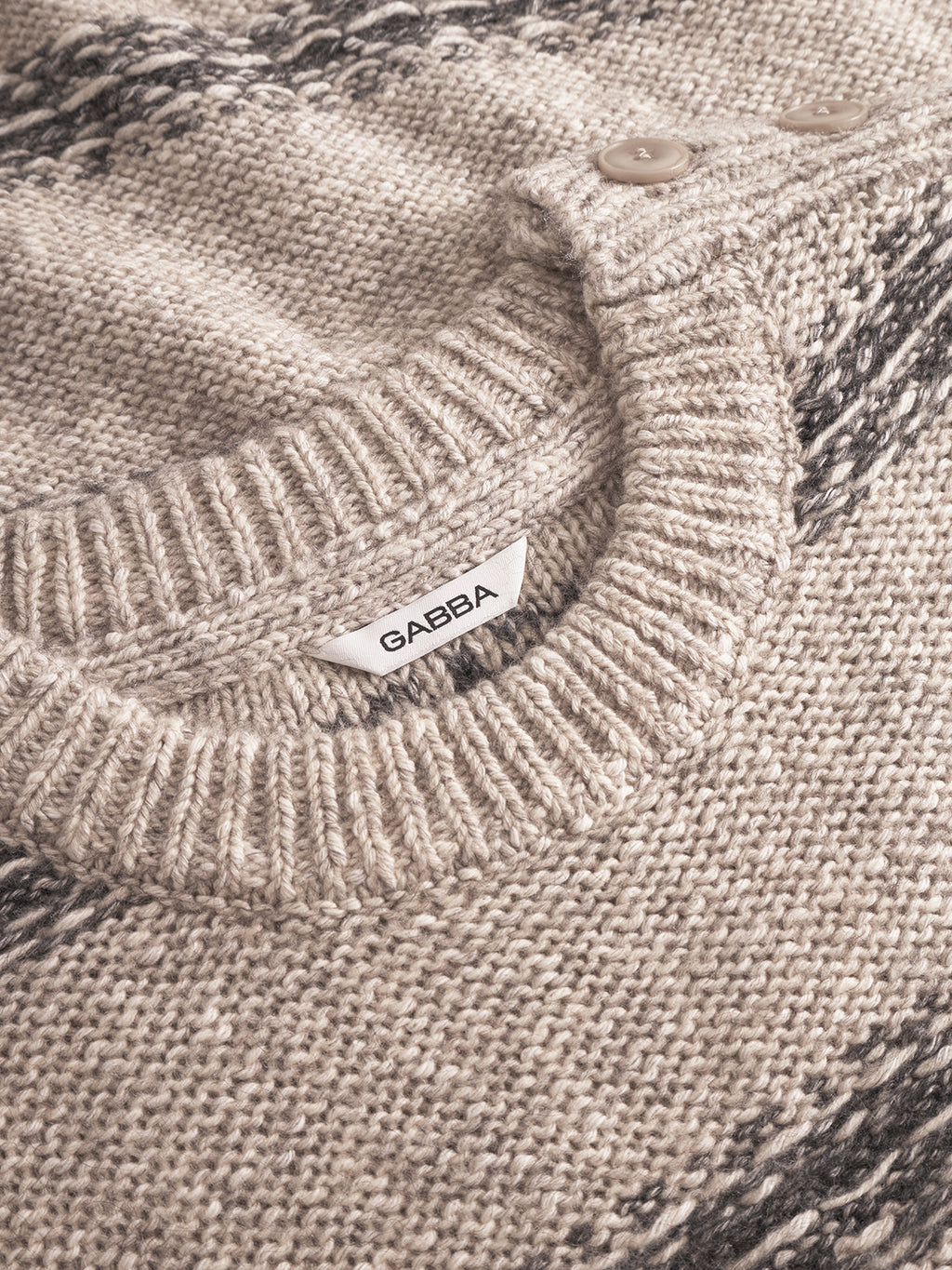 Close-up of the Osaka Fay LS - Ecru by Gabba, a beige and gray melange knit sweater with ribbed neckline, two beige buttons on the shoulder, and a white fabric label showing the Gabba brand name.