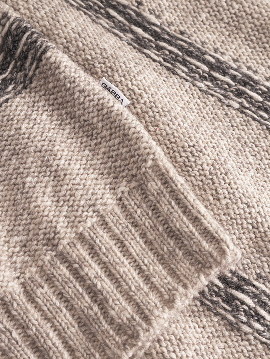 A close-up of the Gabba Osaka Fay LS - Ecru sweater, featuring beige melange yarns with black marbled patterns, ribbed cuffs, and a white Gabba tag.