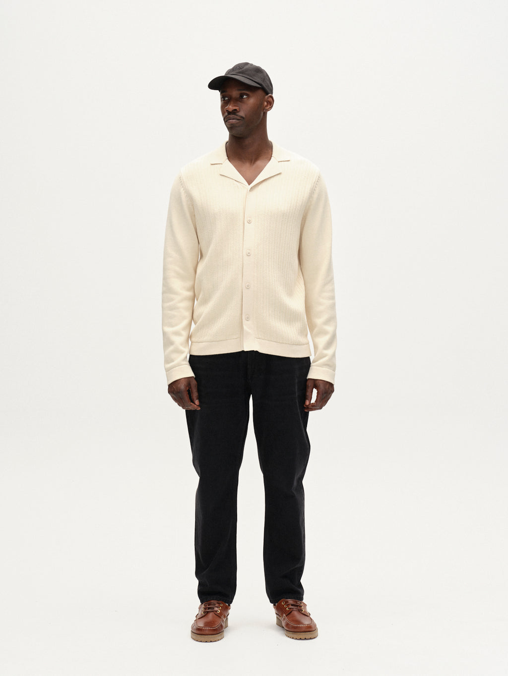 A man stands facing forward against a plain white background, wearing Gabbas Sakai Wong LS cardigan in White Almond, paired with black pants, brown shoes, and a black cap.
