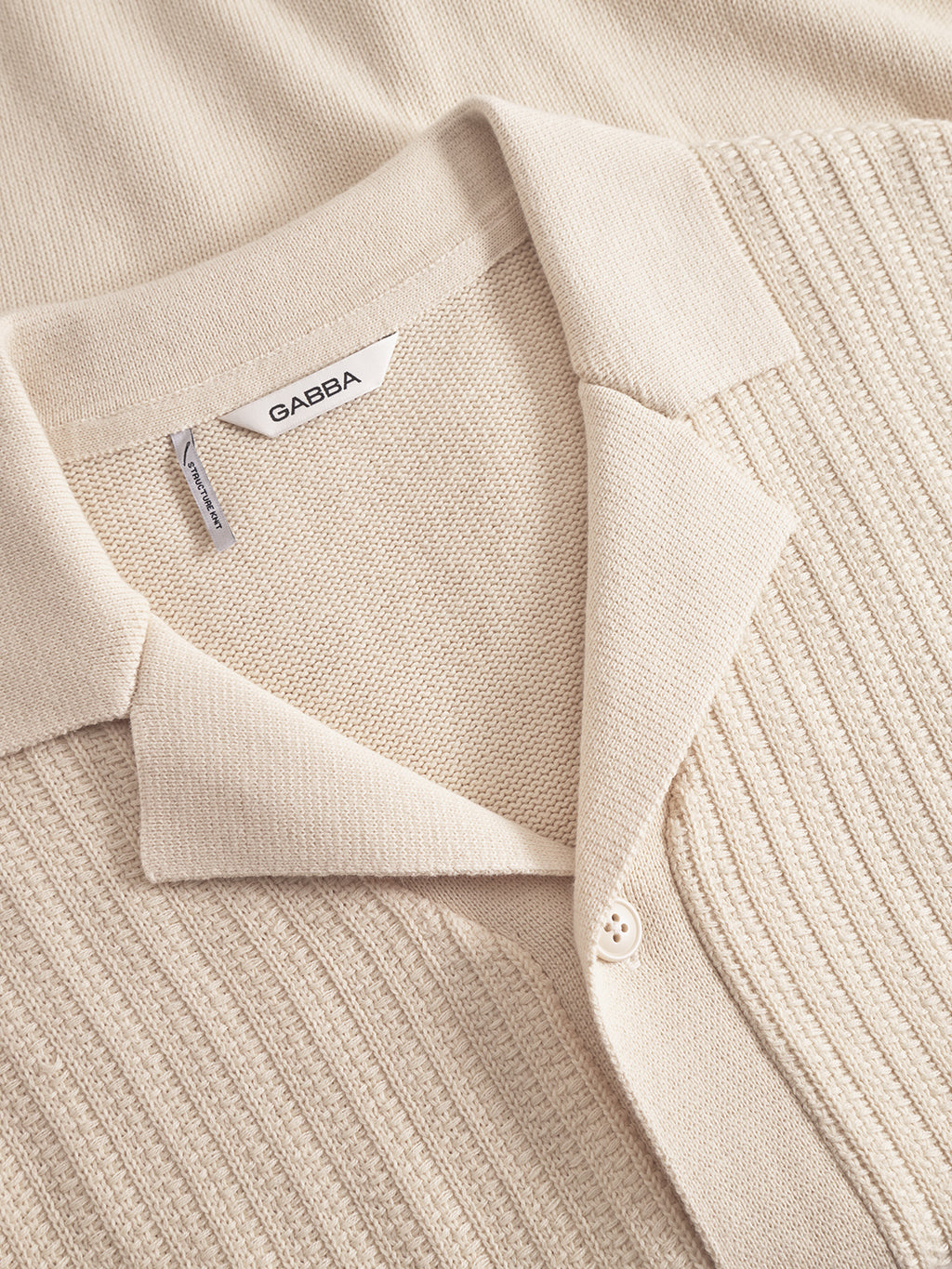 A close-up of the Gabba Sakai Wong LS polo in White Almond, made from 100% cotton with a ribbed front, two buttons, pointed collar, and Gabba tag. The shirt is laid flat on a matching beige surface.