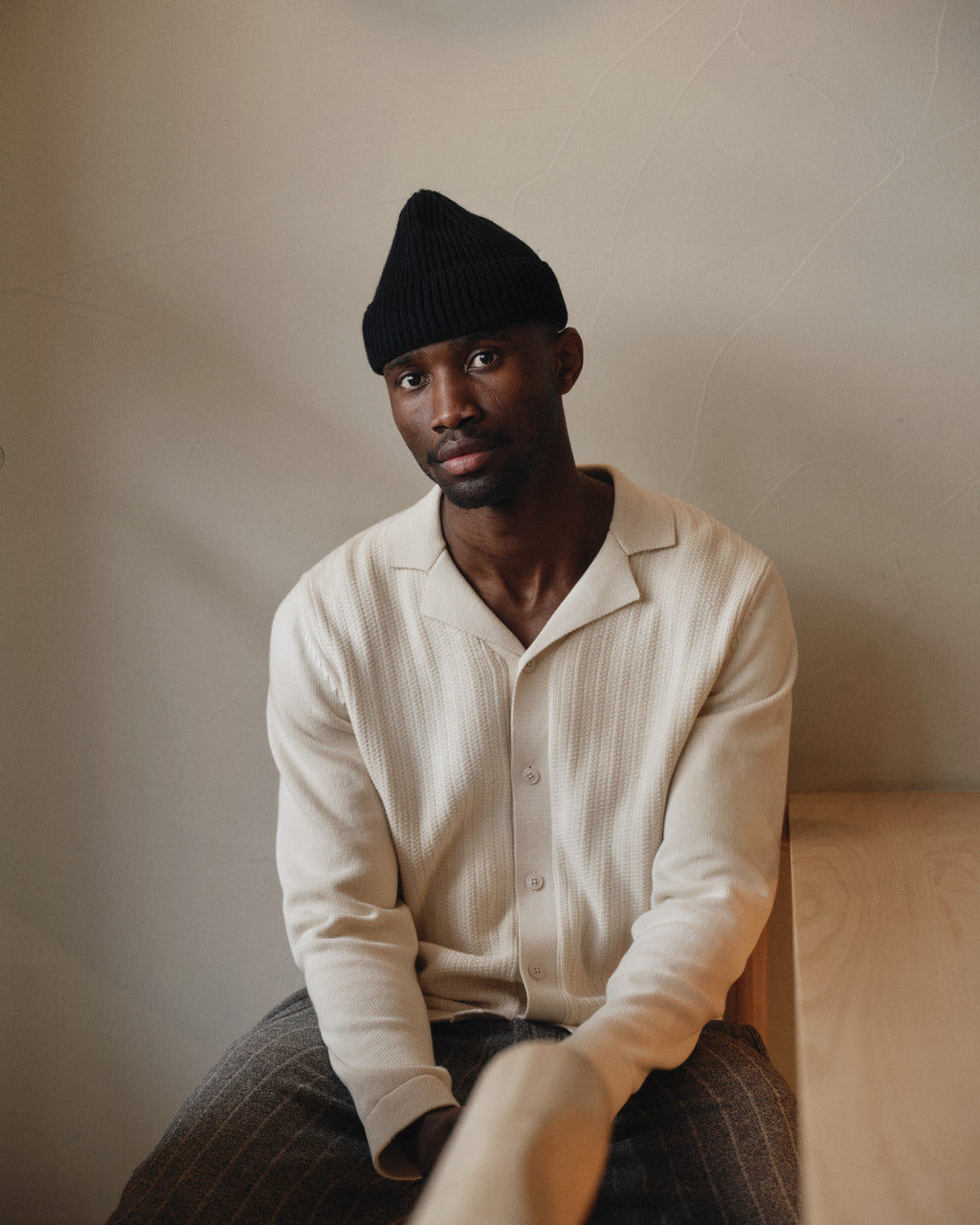 A man wears the Gabba Sakai Wong LS - White Almond shirt, paired with gray pants and a black knit beanie, as he sits calmly on a wooden bench indoors against a neutral wall.