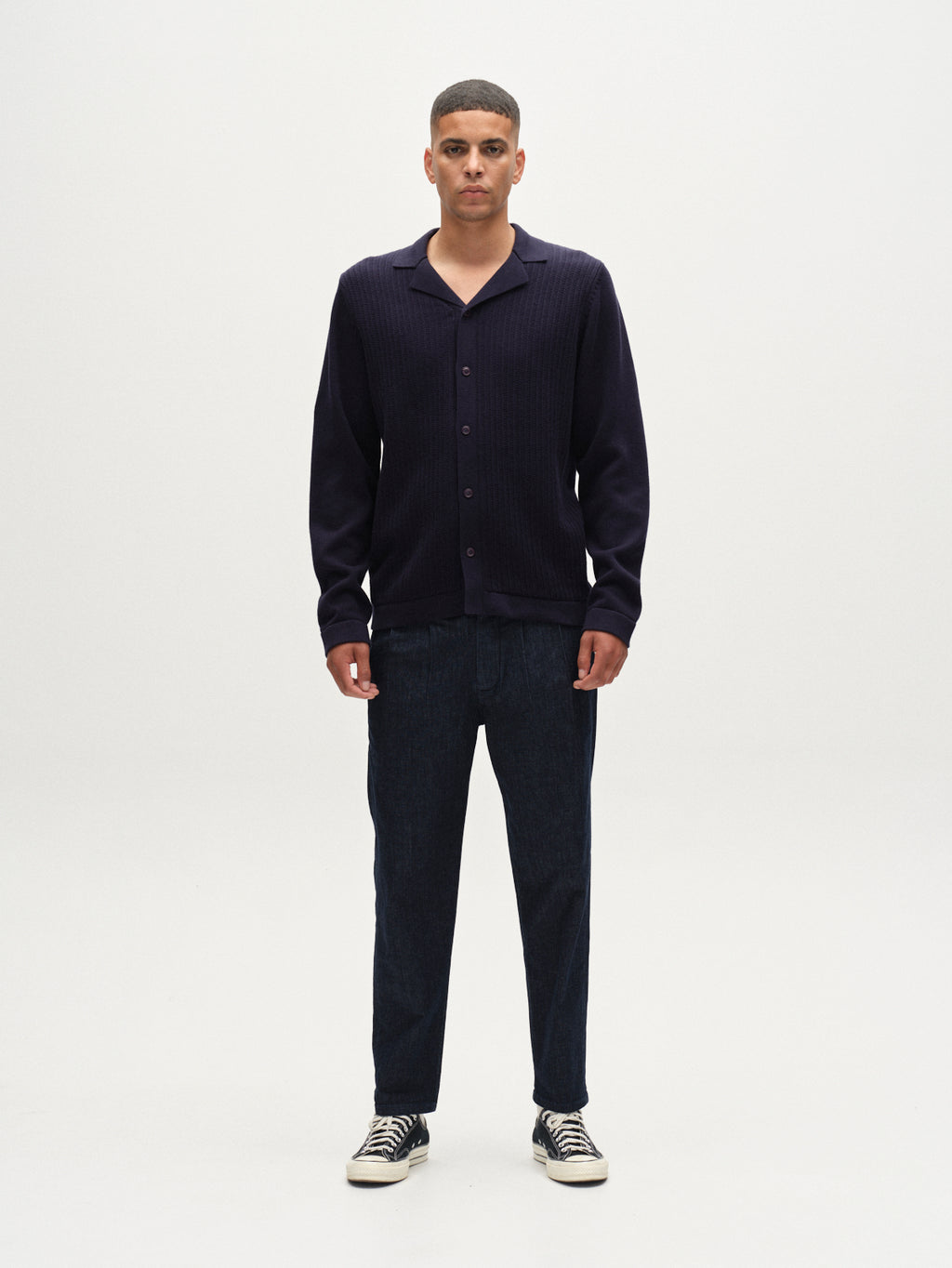 A man stands facing forward against a plain white background, wearing the Gabba Sakai Wong LS cardigan in navy, dark pants, and black-and-white sneakers.