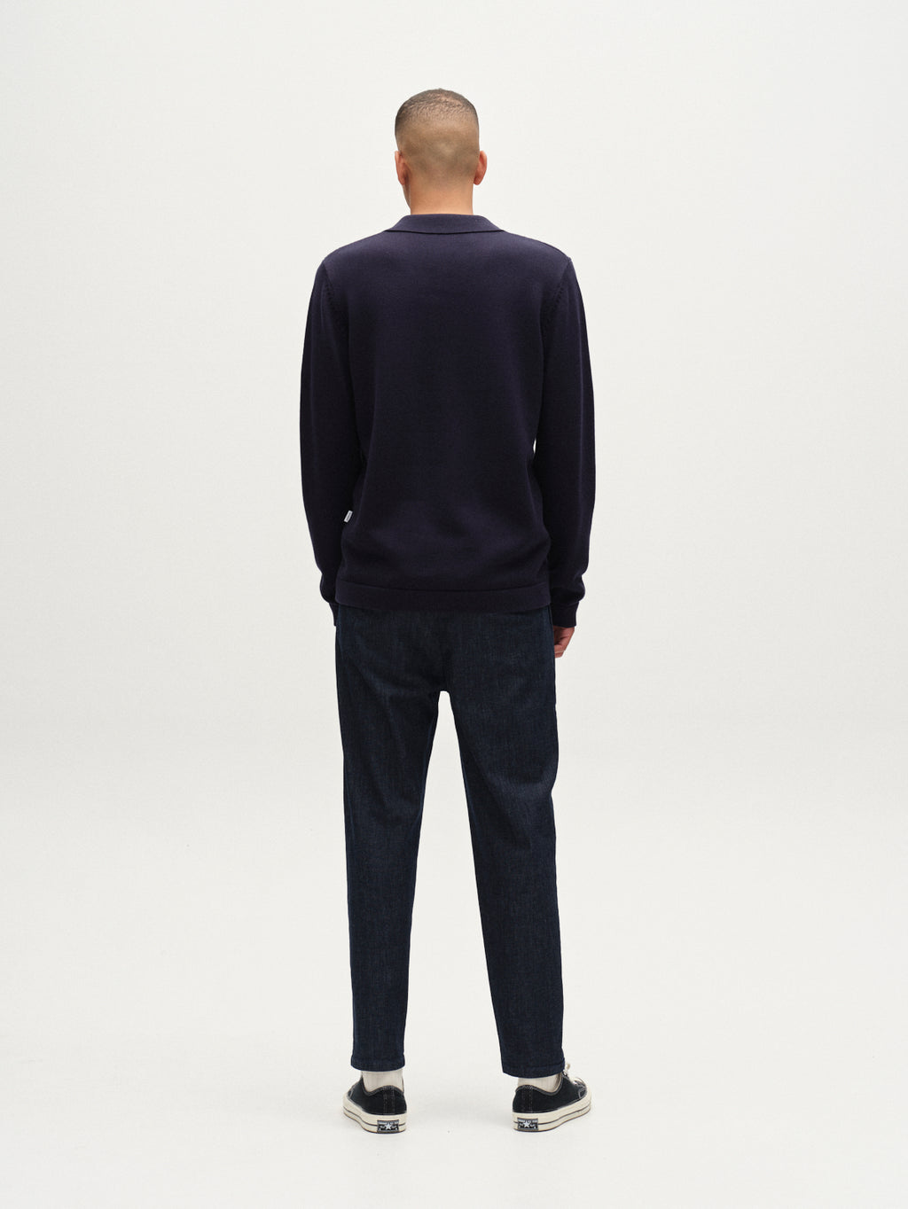 A person stands with their back to the camera against a plain white background, wearing the Gabba Sakai Wong LS - Navy cardigan, dark pants, and black sneakers with white soles.