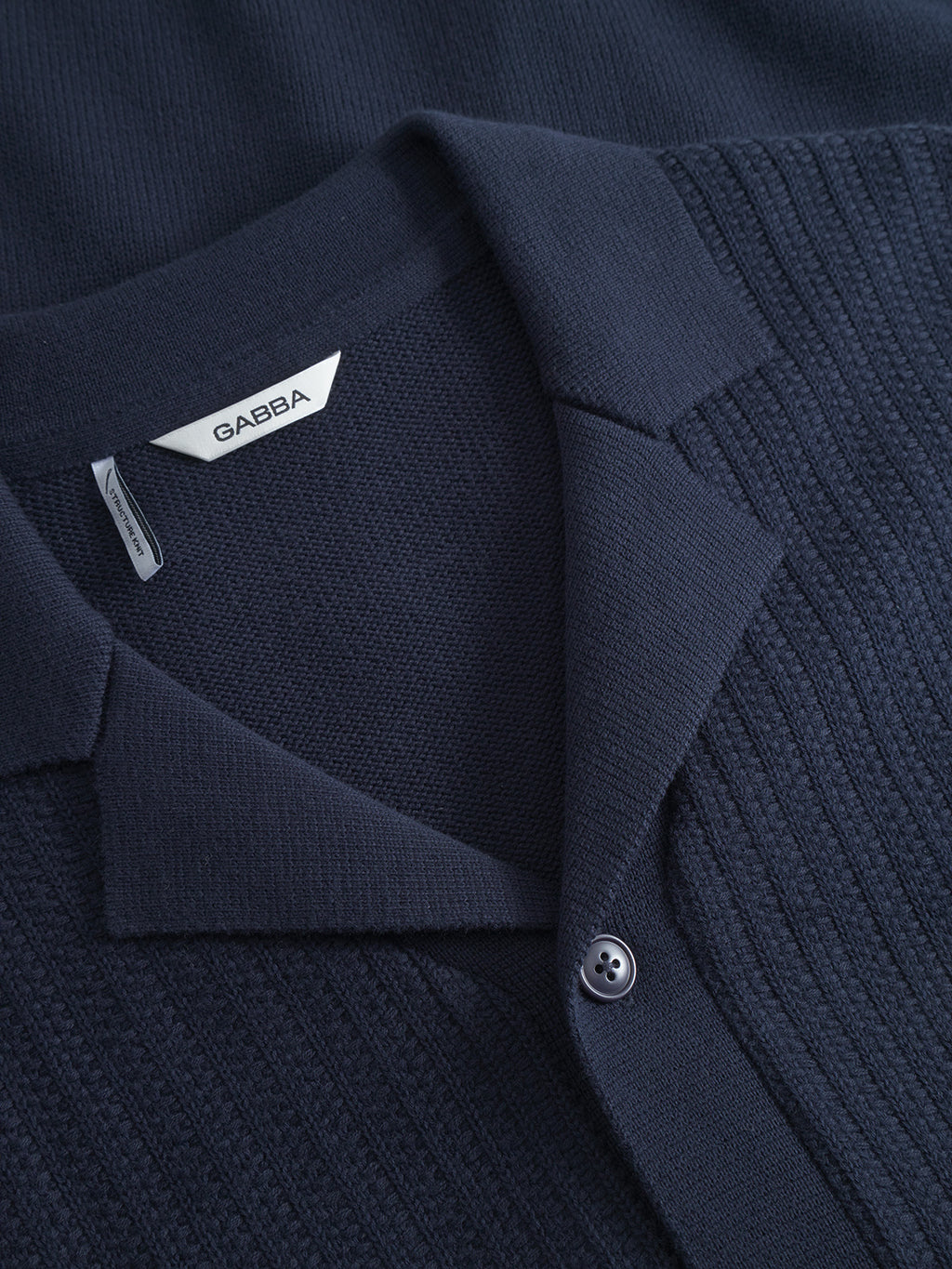 Close-up of the Gabba Sakai Wong LS - Navy shirt, showing its resort collar, single button, and a label reading Gabba. The navy cotton knit features both ribbed and smooth textures for added detail.