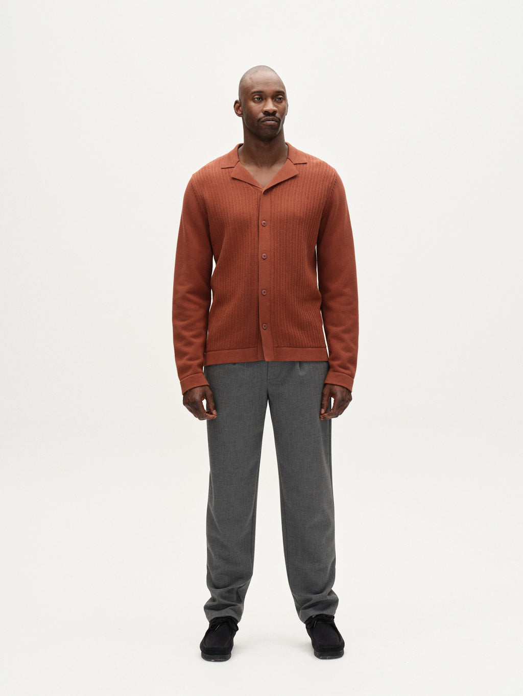 A man faces forward against a plain white background, wearing the Gabba Sakai Wong LS cardigan in brown rust, paired with gray pants and black shoes.