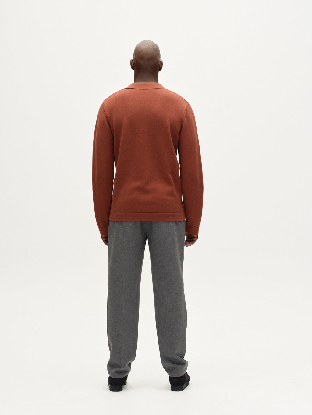 A person stands facing away from the camera against a plain white background, wearing the Gabba Sakai Wong LS cardigan in brown rust with a resort-style collar, paired with gray pants.