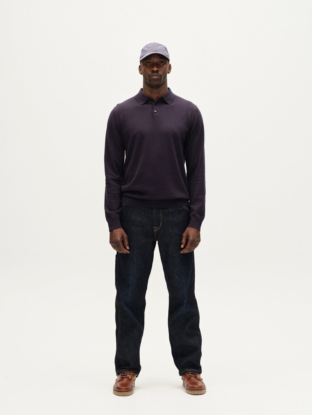 A man stands facing forward against a plain white background wearing the Gabba Akita Prex LS - Navy, a modern wardrobe essential, paired with dark blue jeans, a light purple cap, and brown shoes for a stylish look.