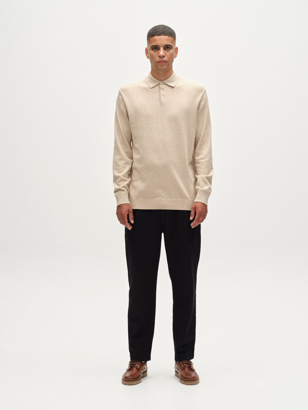 A man faces forward against a plain white background, wearing the Gabba Akita Prex LS - Sand long-sleeve polo shirt, black pants, and brown shoes for a look of refined comfort.