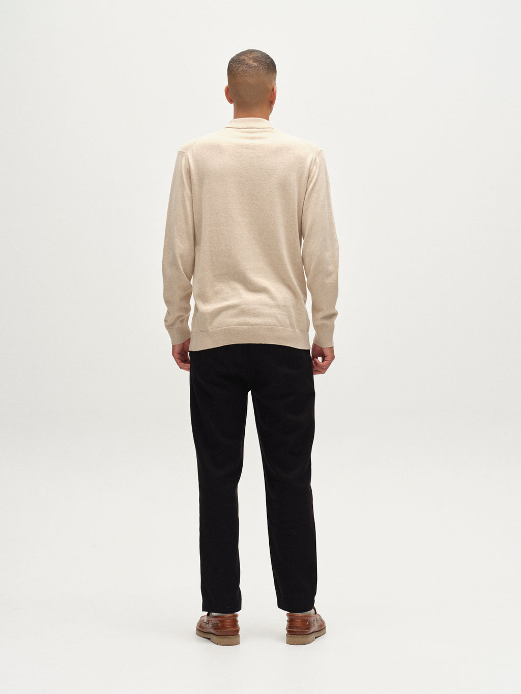 A person stands facing away from the camera, wearing the Gabba Akita Prex LS - Sand long-sleeve polo with black pants and brown shoes, set against a plain white background.