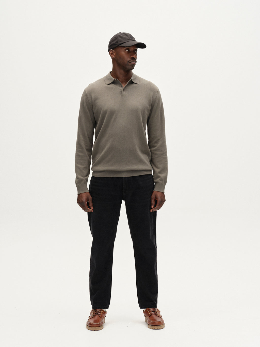 A man stands against a white background wearing the Gabba Akita Prex LS long-sleeve polo in Dust Sage Green, paired with black pants, brown shoes, and a dark cap. He faces forward with hands by his sides, looking slightly left.