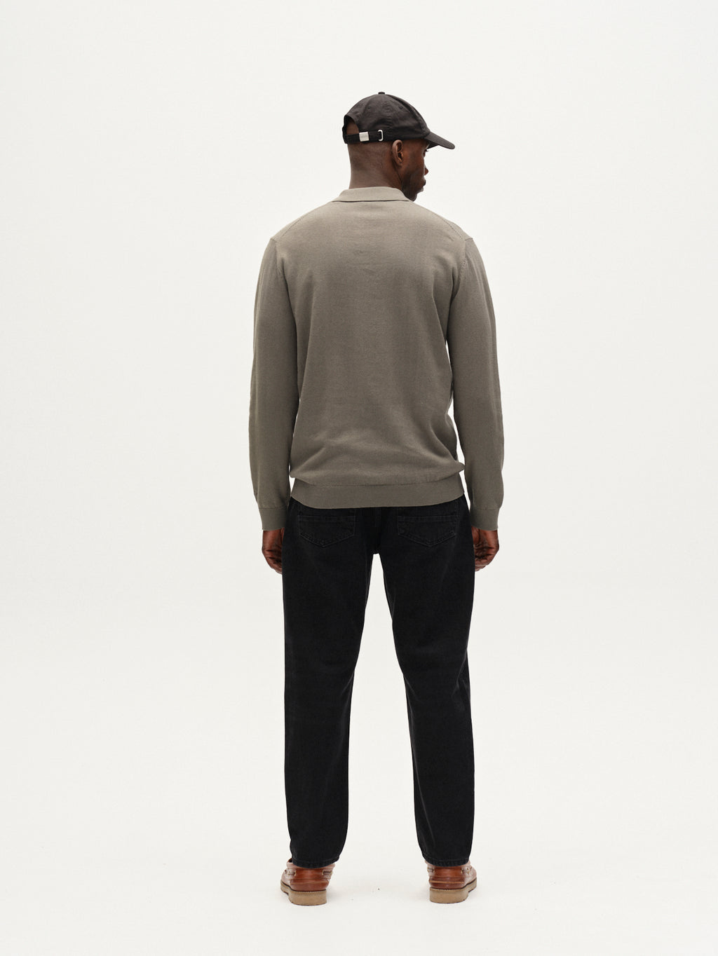 A man faces away from the camera, wearing Gabbas Akita Prex LS in Dust Sage Green with black pants, brown shoes, and a black baseball cap, set against a plain white background.