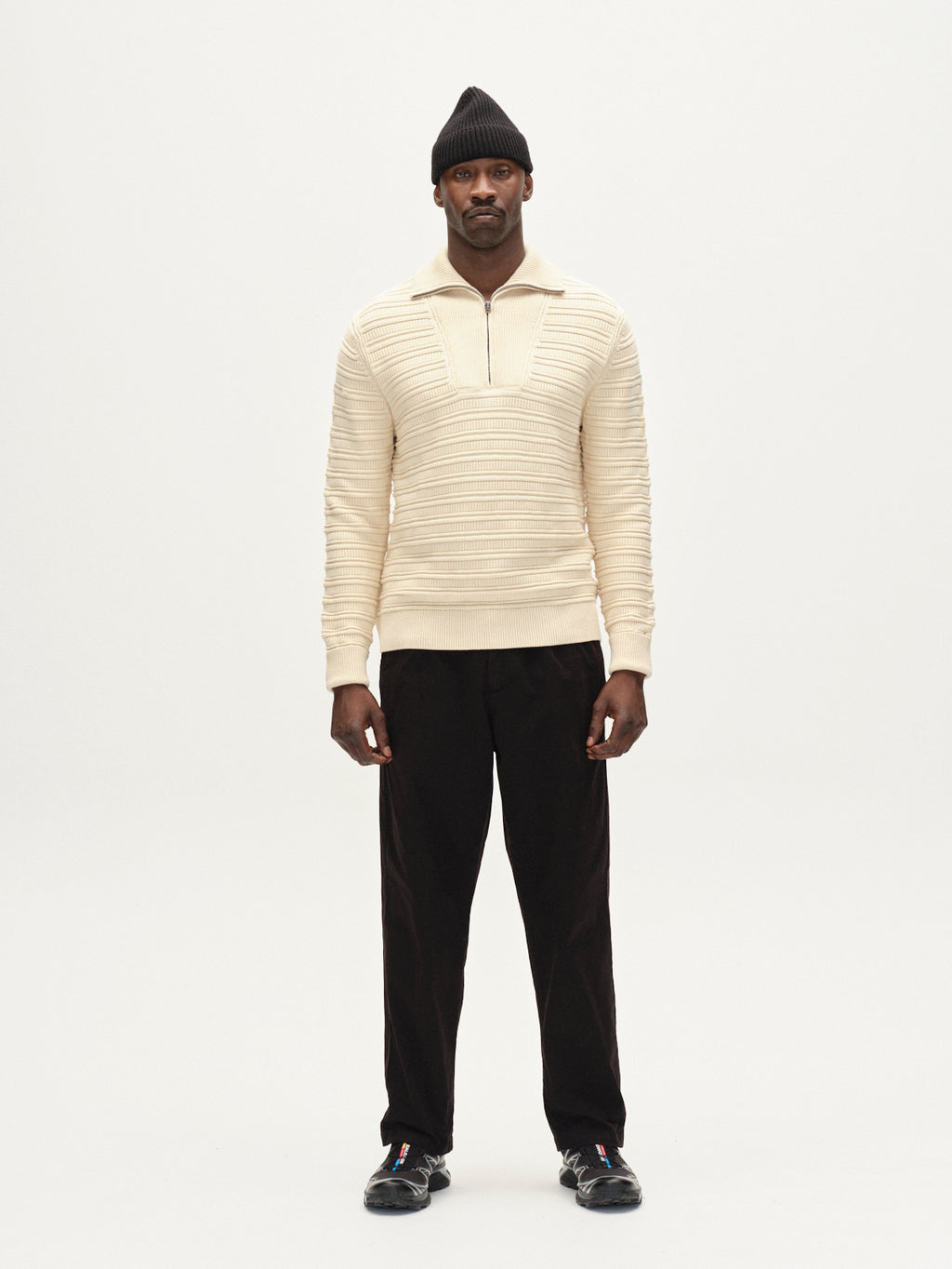 A man faces forward wearing the Gabba Iwaki Buco LS - White Almond zip jumper, featuring a textured pattern and ribbed collar, paired with black pants, sneakers, and beanie against a plain white background.