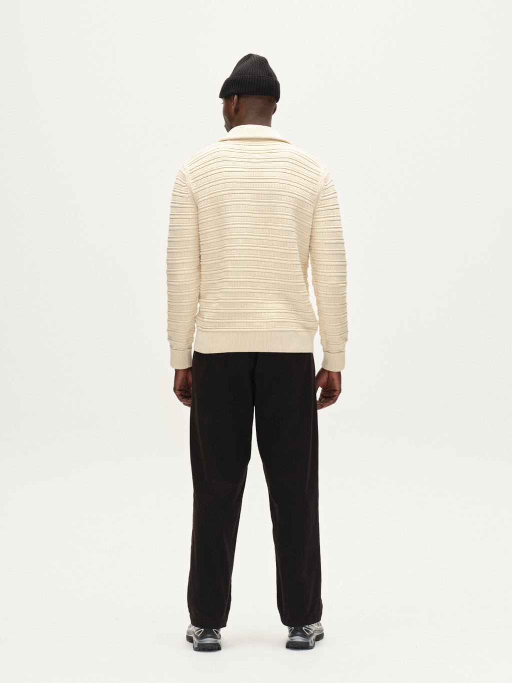 A man faces away, wearing the Gabba Iwaki Buco LS - White Almond jumper with a textured pattern, black pants, black beanie, and dark sneakers. The background is plain and light-colored.