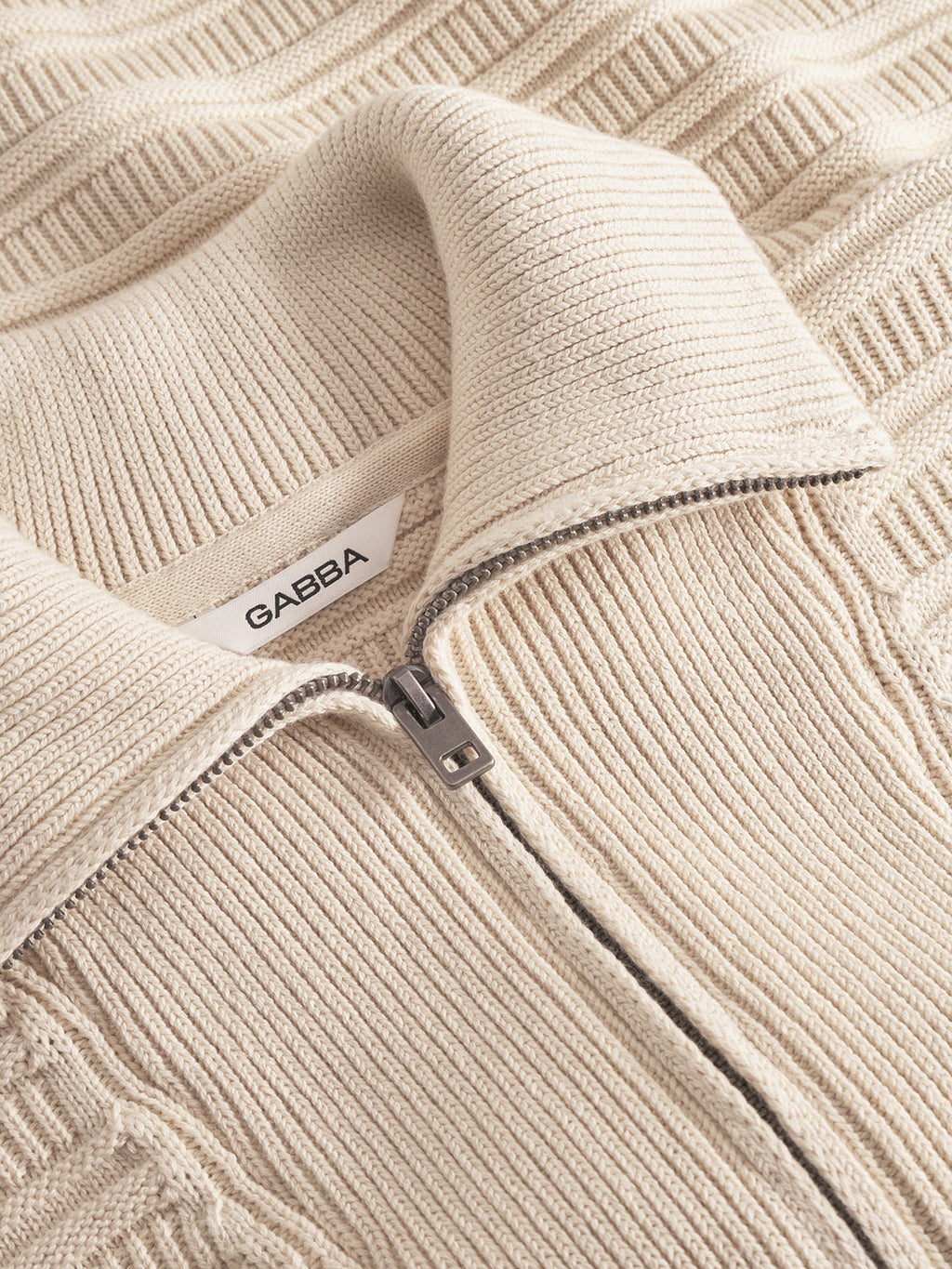 Close-up of the Iwaki Buco LS jumper by Gabba in White Almond. Features a textured beige cotton fabric, ribbed half-zip collar with metal zipper, and a white “Gabba” label inside the folded collar.