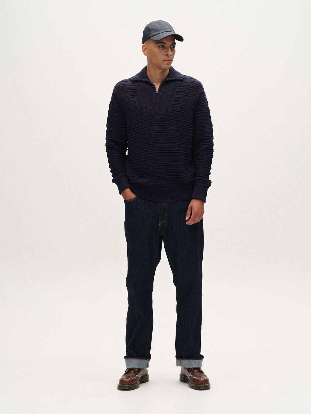 A man stands in a studio against a white background, wearing the Gabba Iwaki Buco LS - Navy sweater, paired with dark jeans, brown shoes, and a dark baseball cap. He faces forward with his hands at his sides.