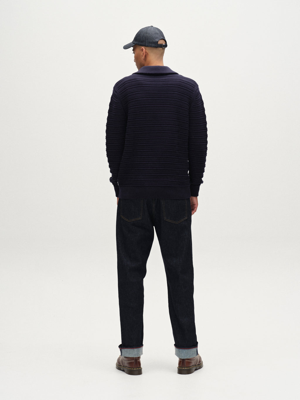 A man faces away from the camera wearing the Gabba Iwaki Buco LS - Navy sweater, paired with a dark baseball cap, cuffed jeans, and brown shoes against a plain light background.