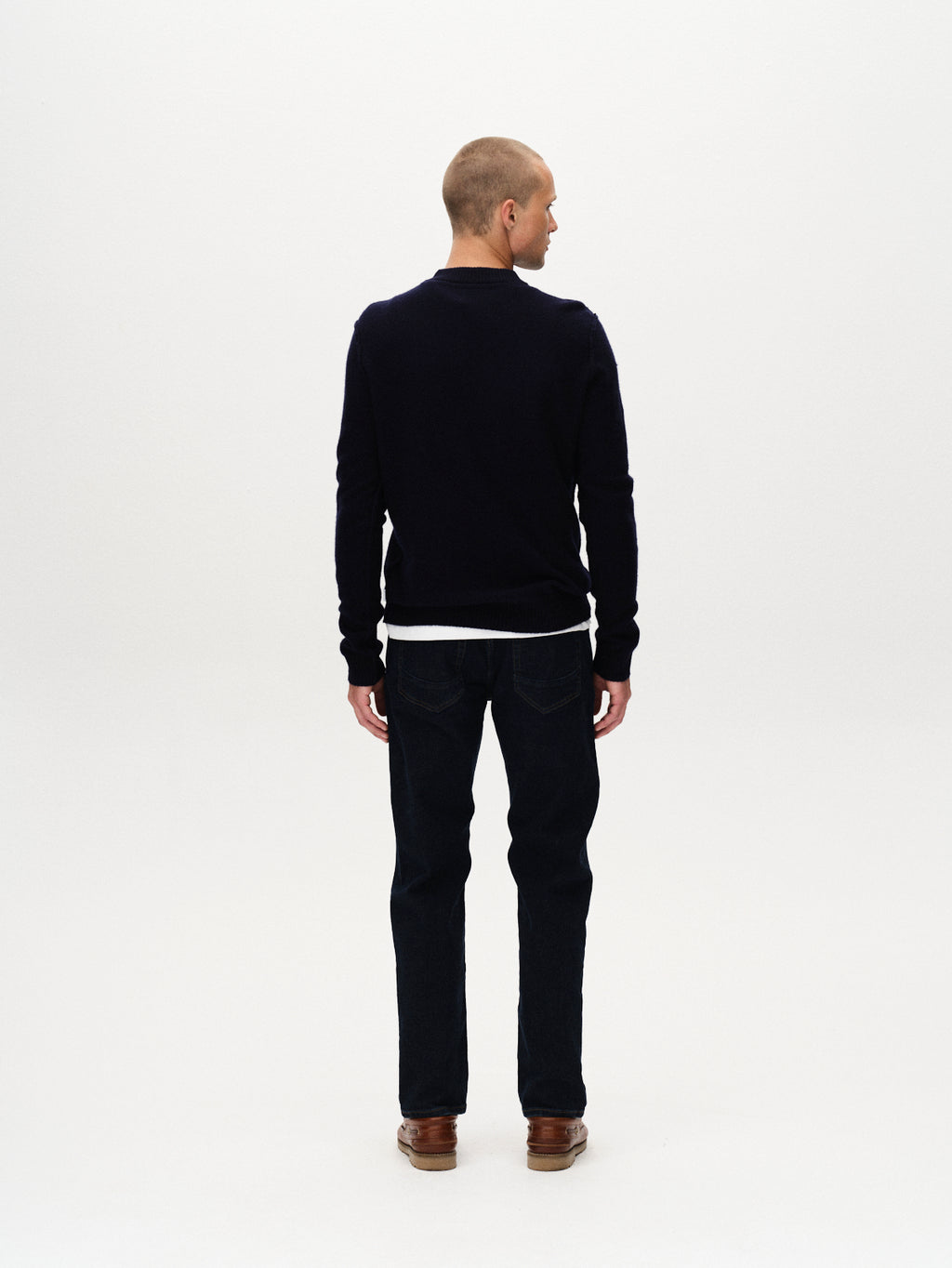 A man with short hair, dressed in a dark sweater, white undershirt, brown shoes, and Gabba’s Marc Jingo - Dark Blue Denim jeans, stands facing away from the camera against a plain white background.