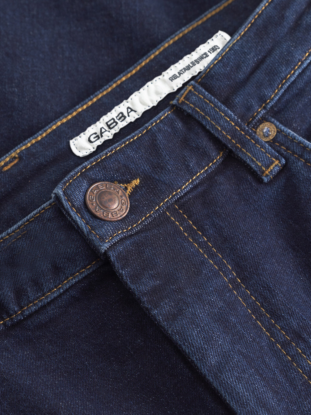 Close-up of Gabba Marc Jingo dark blue denim jeans with a metal button, white Gabba label in the waistband, and visible yellow stitching. These comfort stretch dark rinse jeans offer style and flexibility.