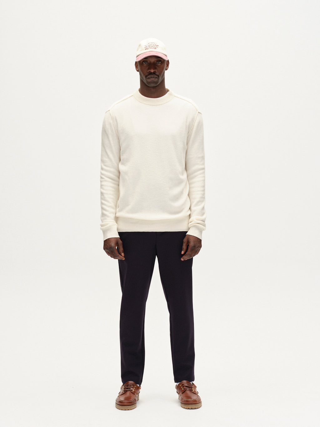 A man stands facing forward against a plain white background, wearing the Gabba Tokota Aico LS in off white, black pants, brown shoes, and a cream baseball cap.