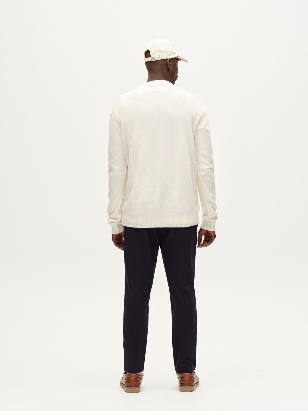 A man stands facing away against a plain white background, wearing the Gabba Tokota Aico LS sweater in off white, paired with black pants, brown shoes, and a cream cap.