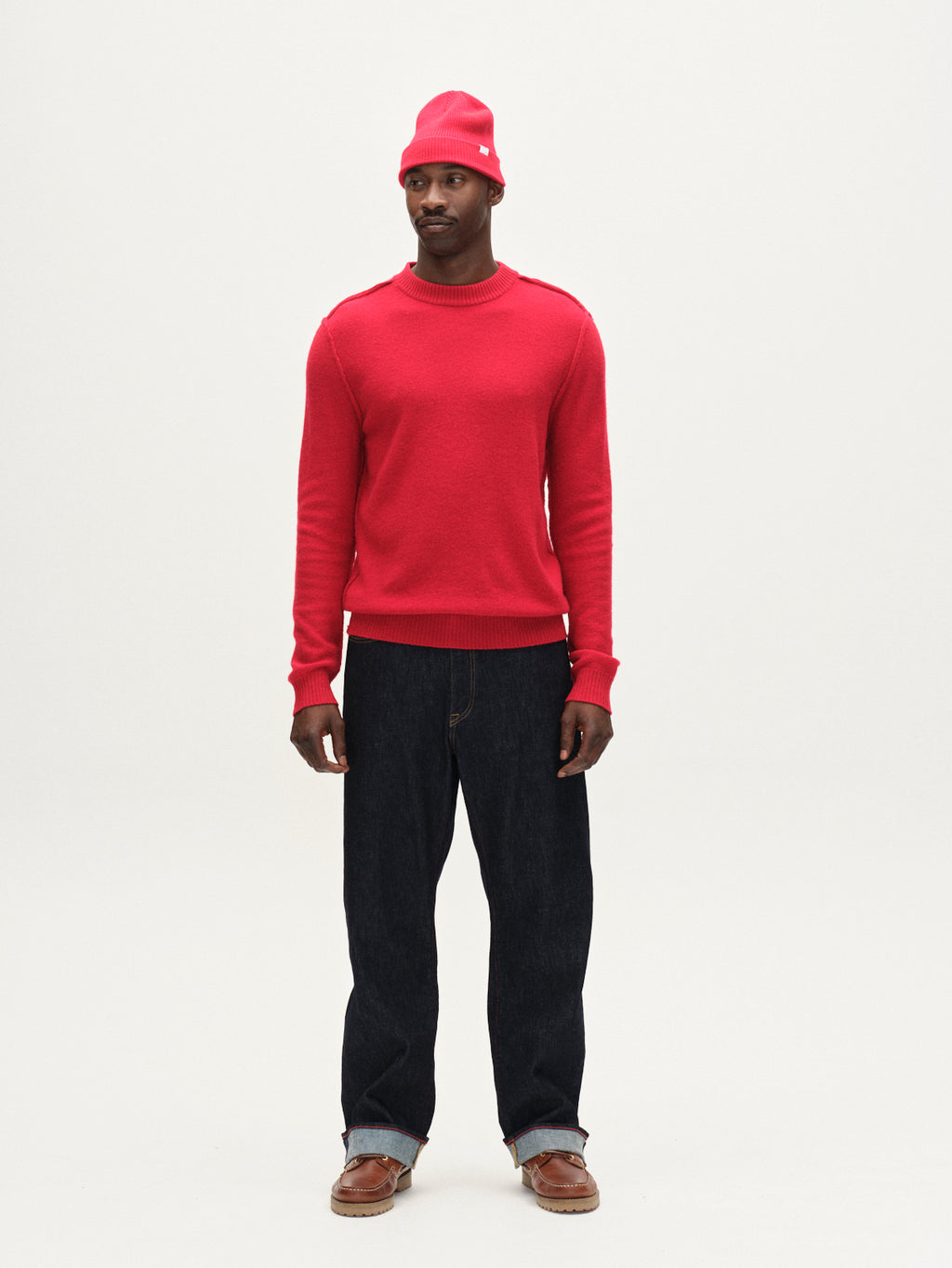 A man faces forward with a neutral expression, wearing the Gabba Tokota Aico LS sweater in red, a matching red beanie, dark blue jeans, and brown shoes against a plain white background.