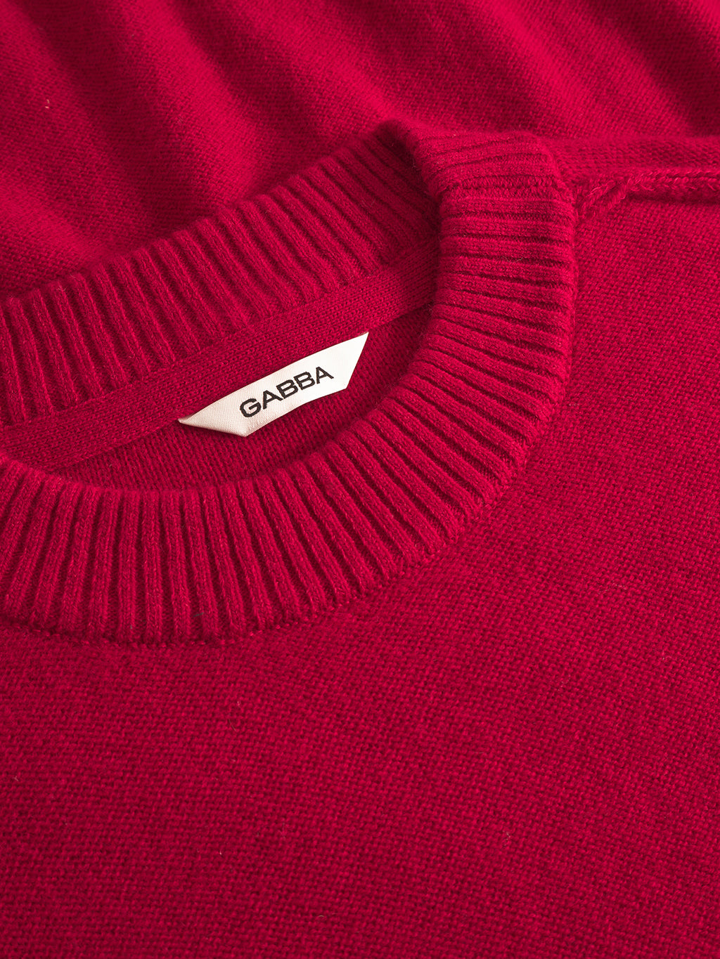 Close-up of the Tokota Aico LS - Red sweater by Gabba, featuring a bright red modern knit with a ribbed crew neckline and a white inner collar label showing the Gabba brand name in black.
