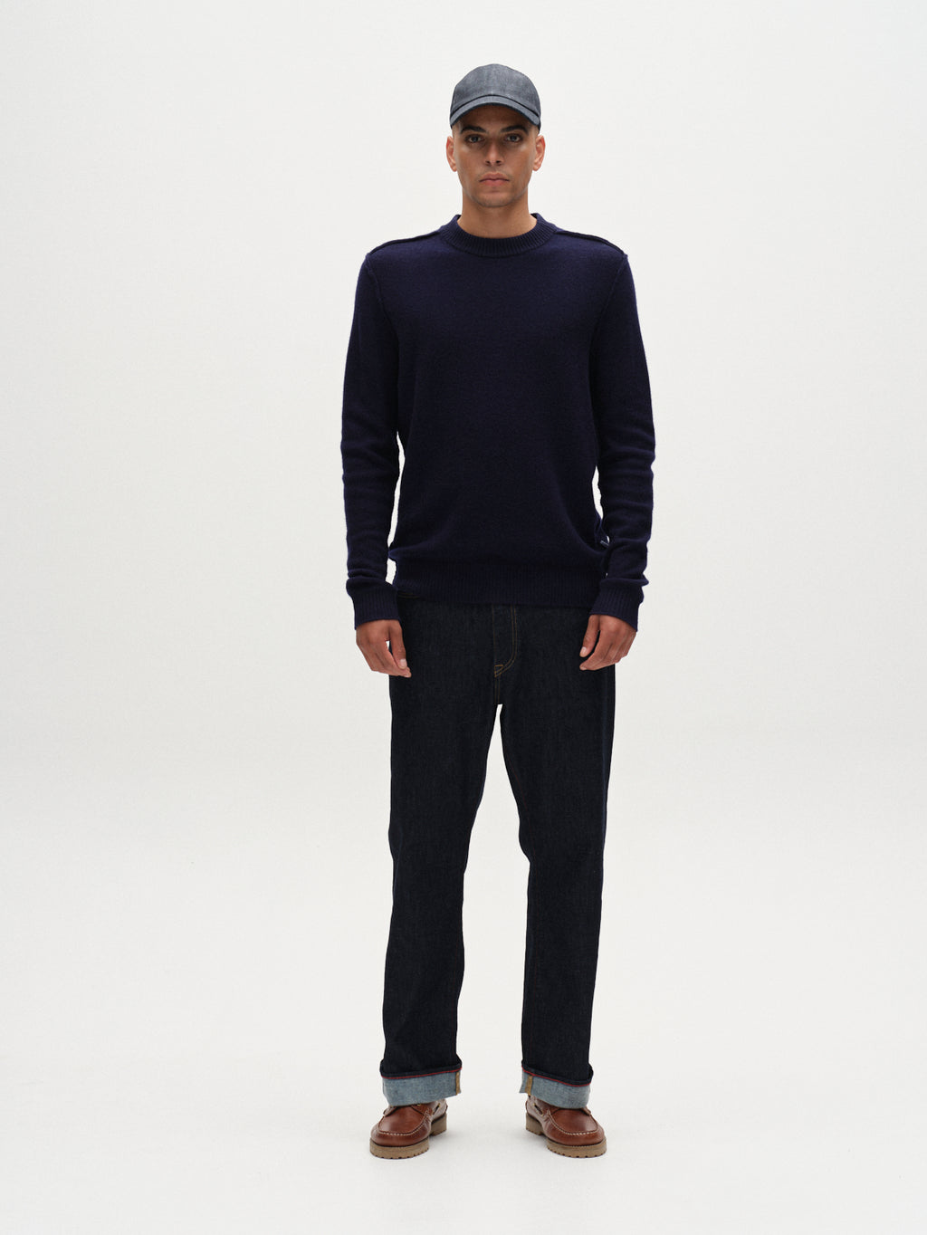 A man stands facing forward against a white background, wearing the Gabba Tokota Aico LS in dark navy—a versatile merino knit sweater—paired with dark jeans, brown shoes, and a gray cap; perfect for any occasion.