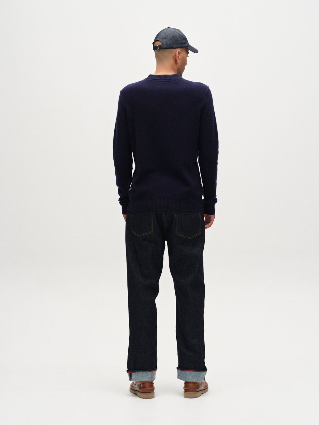 A person faces away from the camera against a plain white background, wearing the Gabba Tokota Aico LS - Dark Navy sweater as a versatile layer, paired with dark cuffed jeans, brown shoes, and a dark baseball cap.