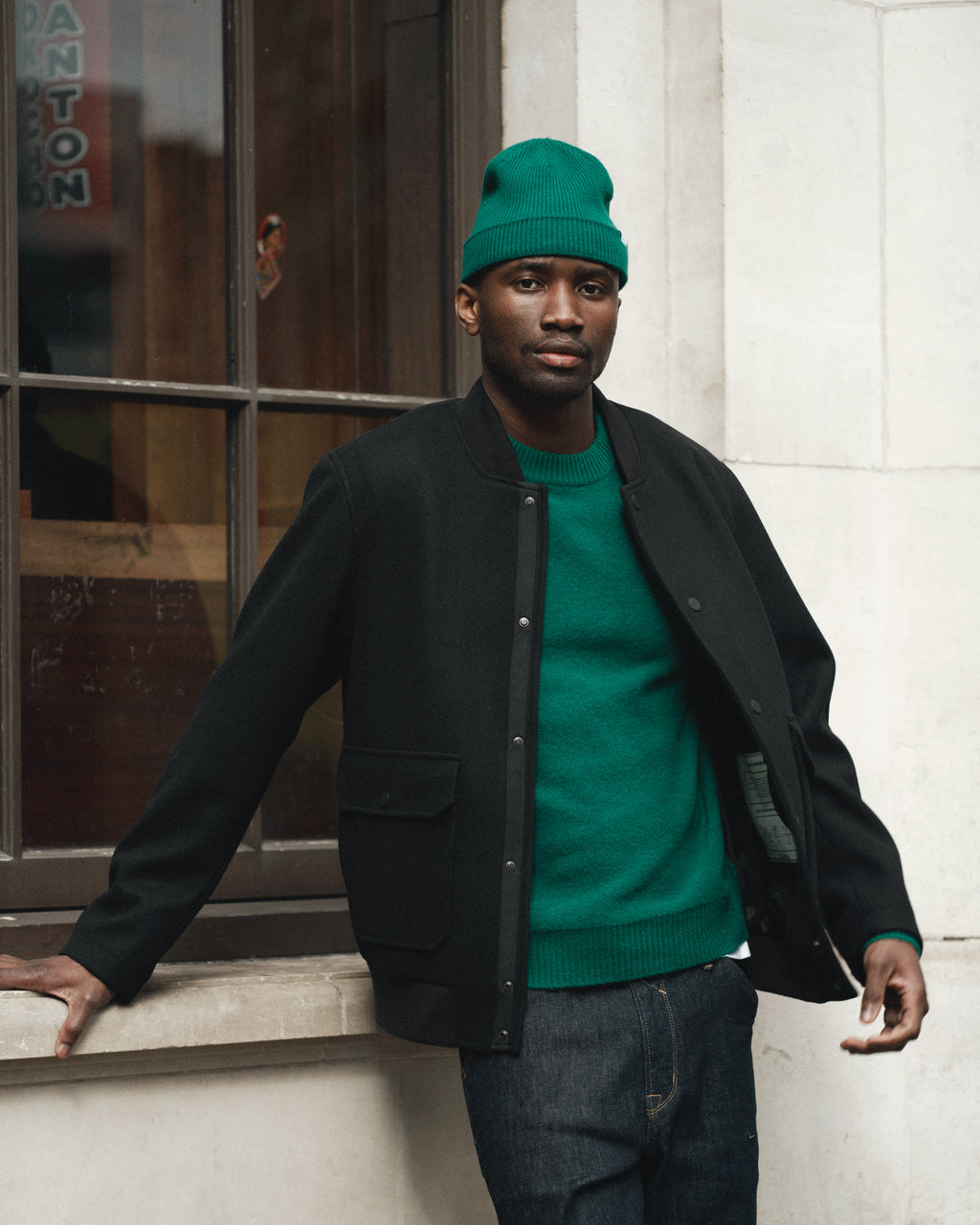 A man in Gabba’s Tokota Aico LS - Green stands by a window outdoors, wearing jeans and a matching green beanie under a black jacket, his hand resting on a stone ledge and his expression neutral.