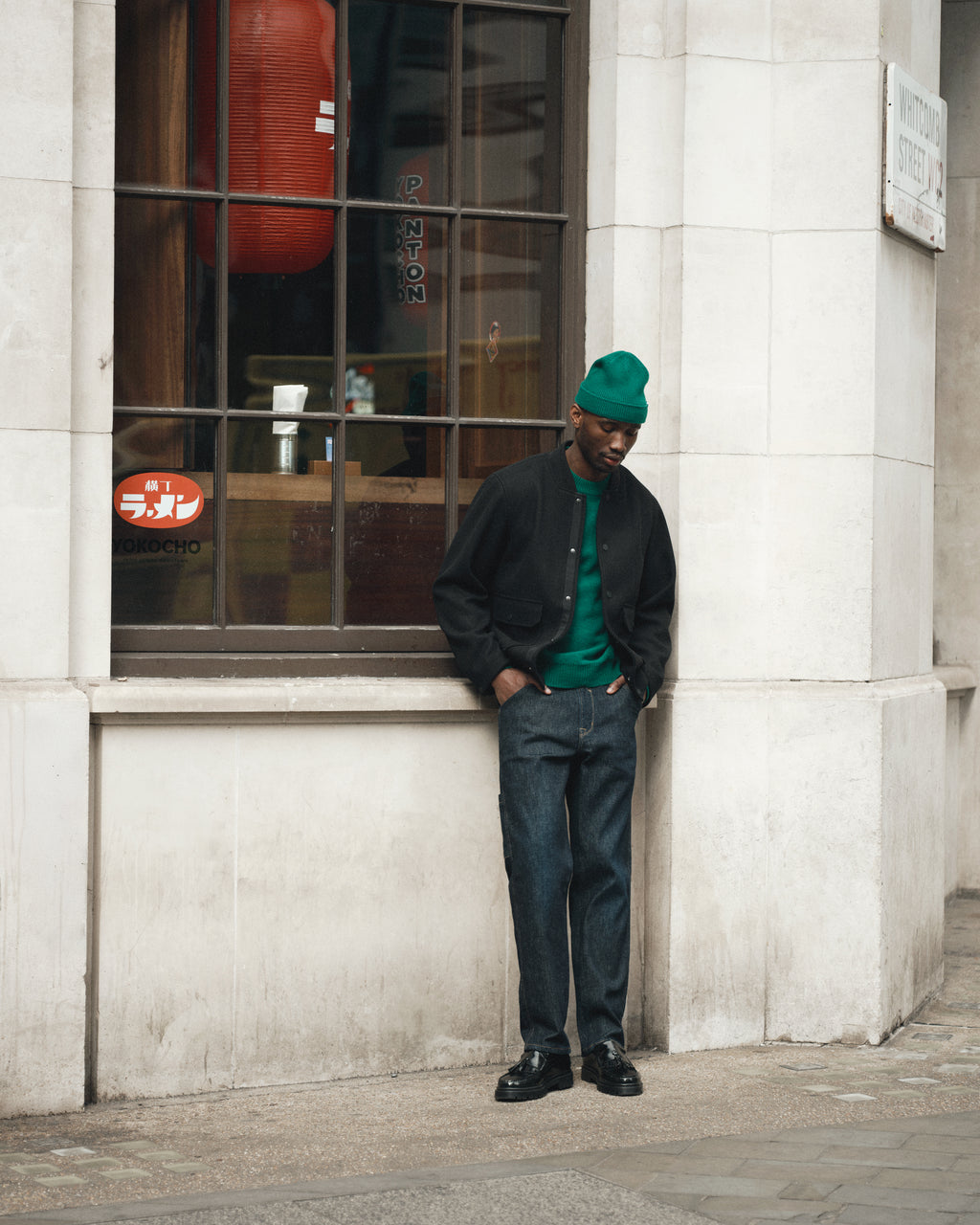 A man in a green Gabba Tokota Aico LS sweater, black jacket, and dark jeans stands on a city sidewalk, leaning against a white stone building with a large window and a red lantern inside.