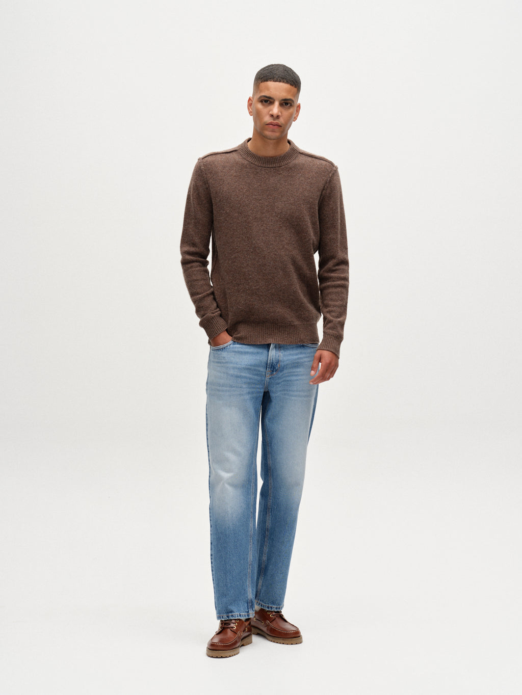 Against a plain white background, a man with short hair and his left hand in his pocket wears the Gabba Tokota Aico LS sweater in dark brown, paired with light blue jeans and brown shoes.