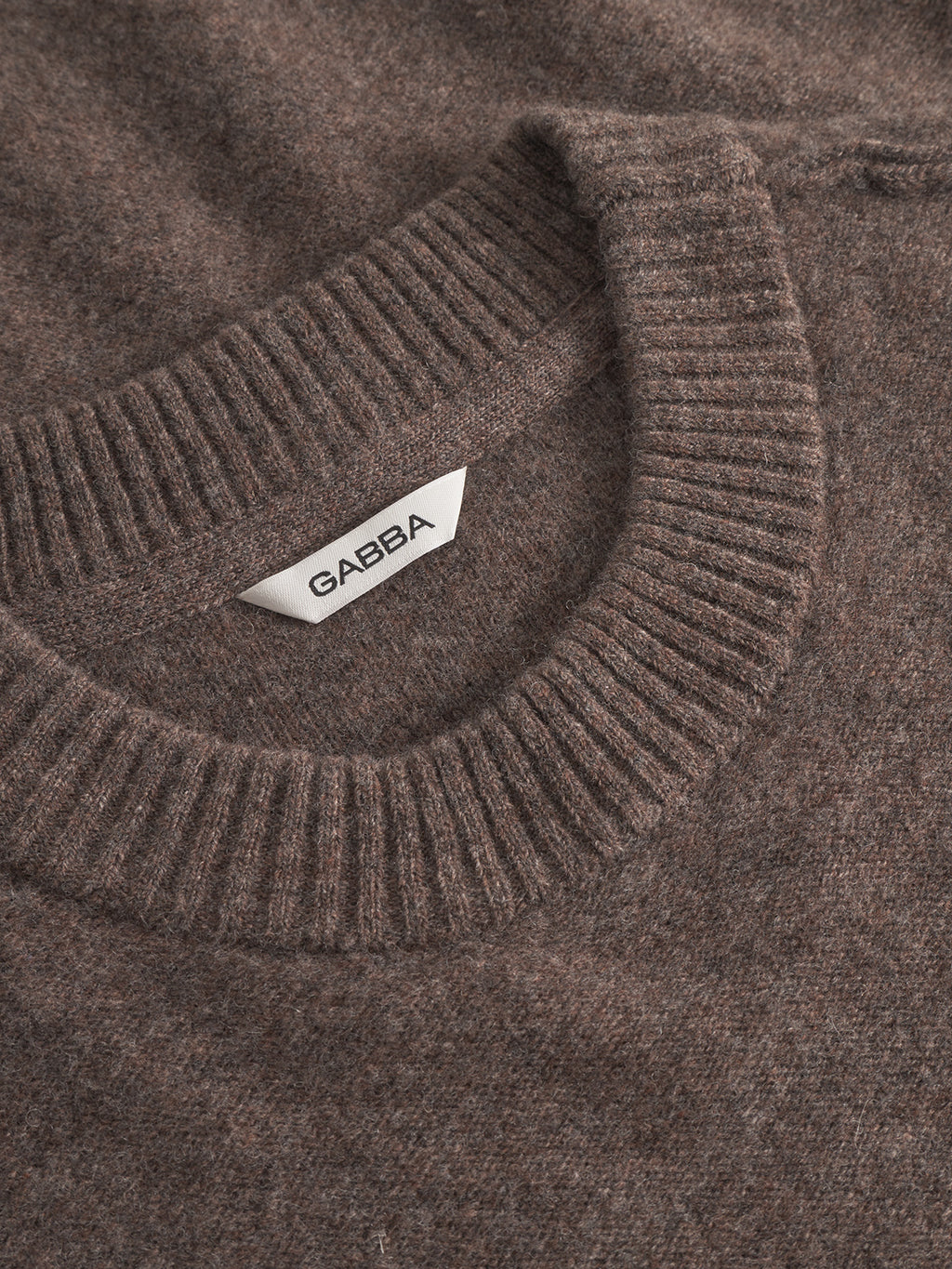 Close-up of the Tokota Aico LS sweater by Gabba in dark brown merino wool, featuring a ribbed crew neck and a white Gabba label inside the neckline.