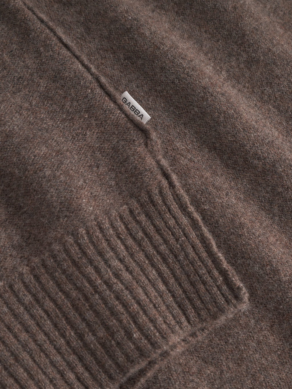 Close-up of the Gabba Tokota Aico LS sweater in dark brown, featuring a visible seam, ribbed cuff, and soft textured knit. A small white fabric tag with Gabba is attached near the seam.