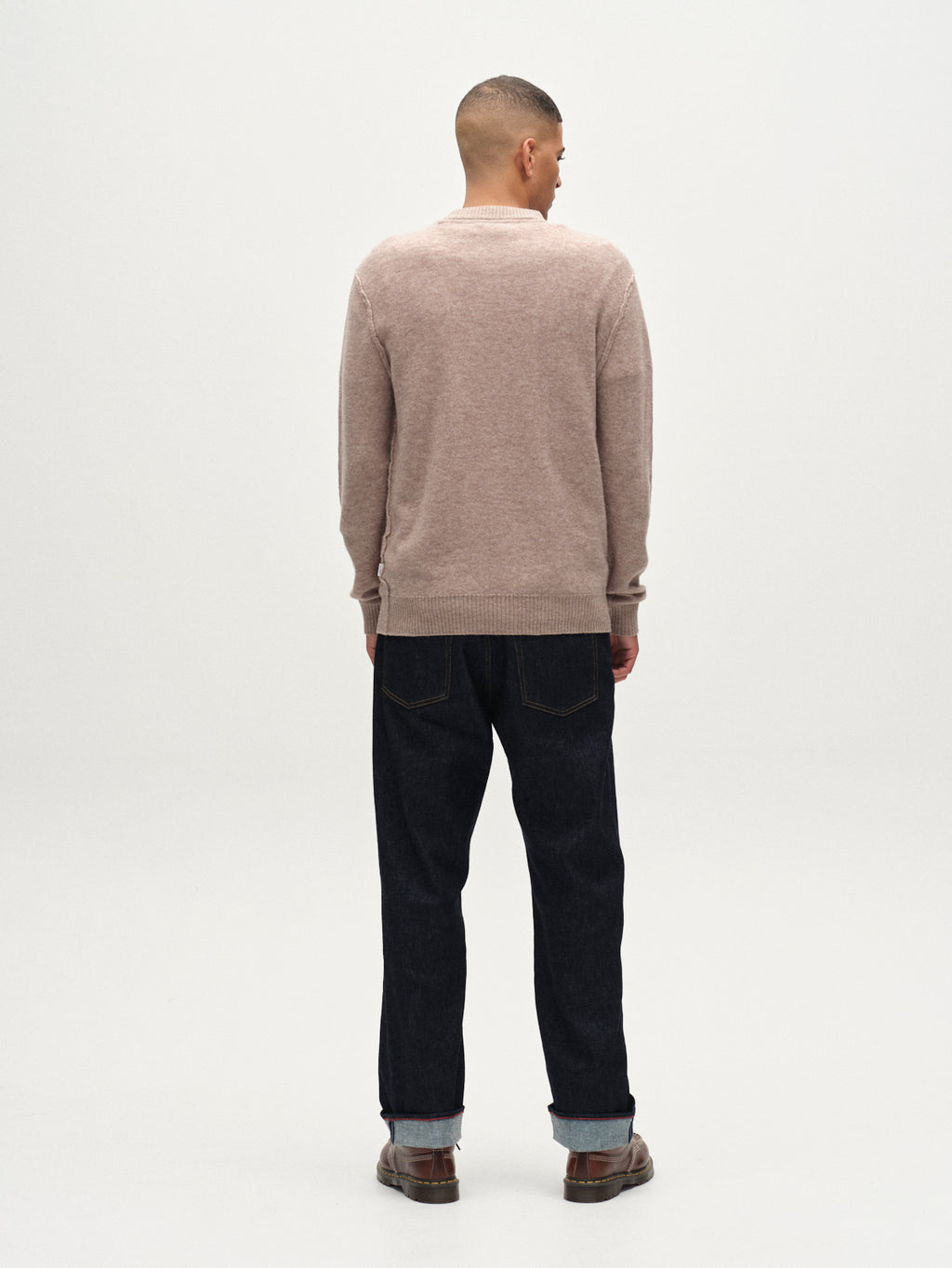 A man with short hair faces away from the camera, wearing the Gabba Tokota Aico LS sweater in light brown, paired with dark jeans and brown shoes against a plain white background.